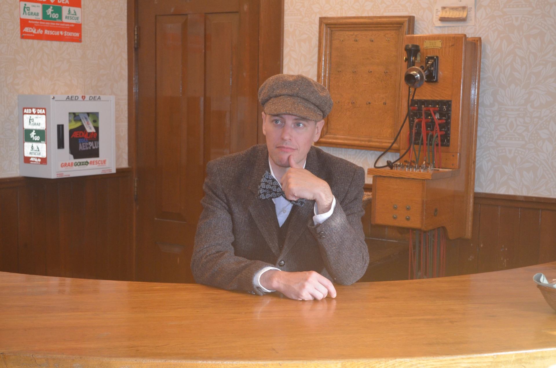 Person in a flat cap sits behind a wooden counter beside an antique switchboard.