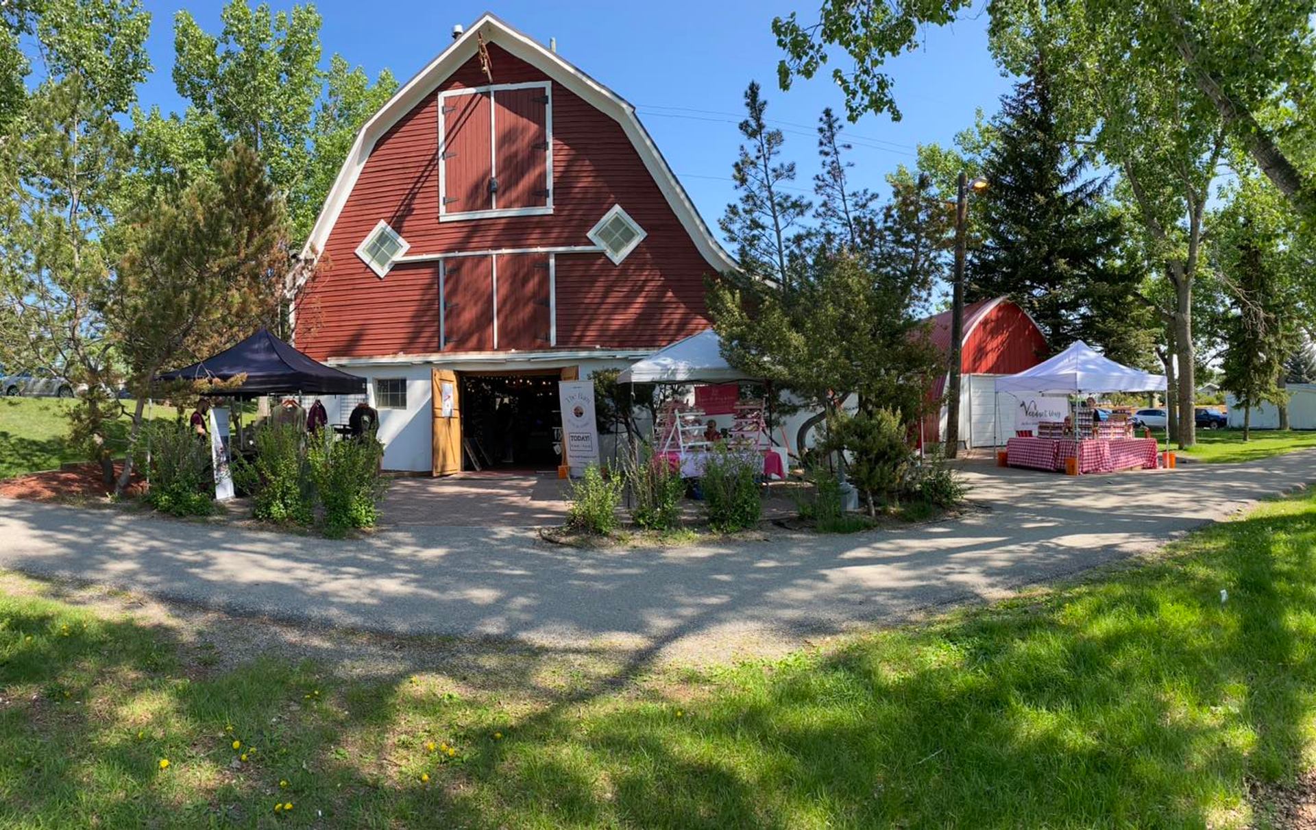 Red barn market with vendor tents, greenery, and visitors on a sunny afternoon.
