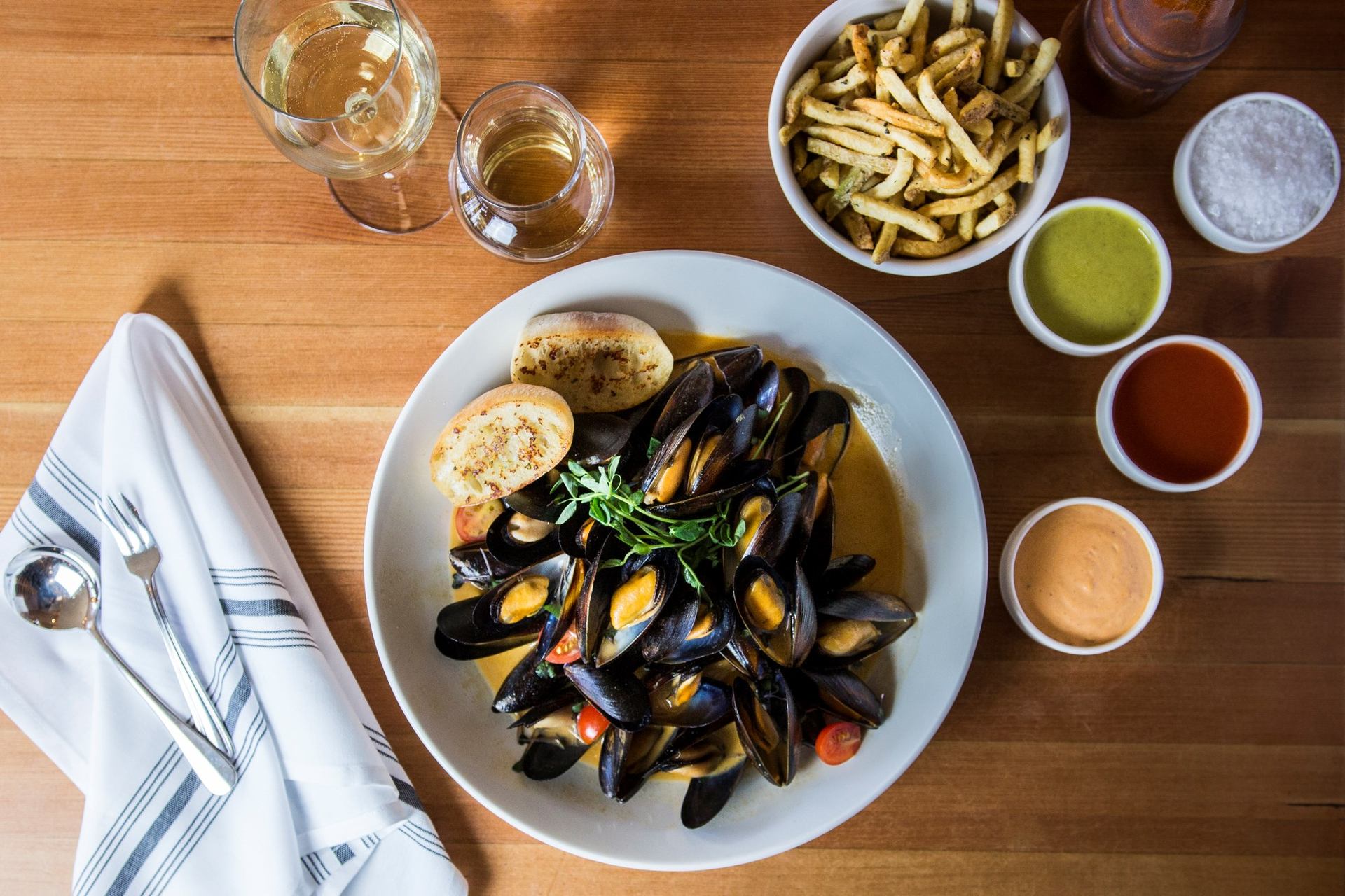 Mussels with herbs, bread, sauces, fries, and wine on a wooden table.