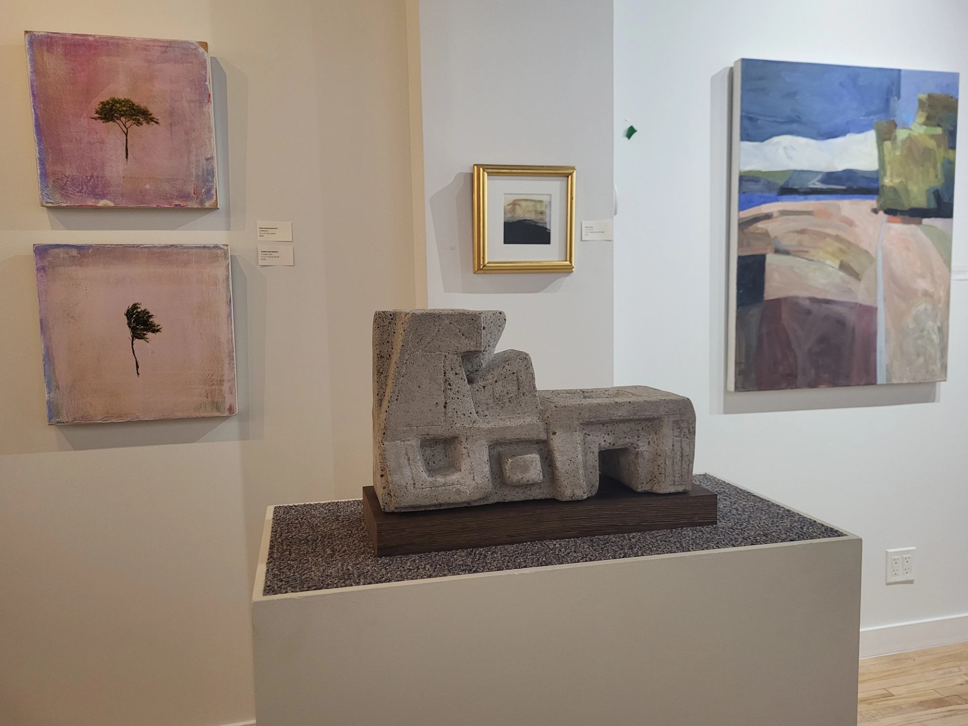 Abstract stone sculpture on a pedestal surrounded by framed paintings in an art gallery.