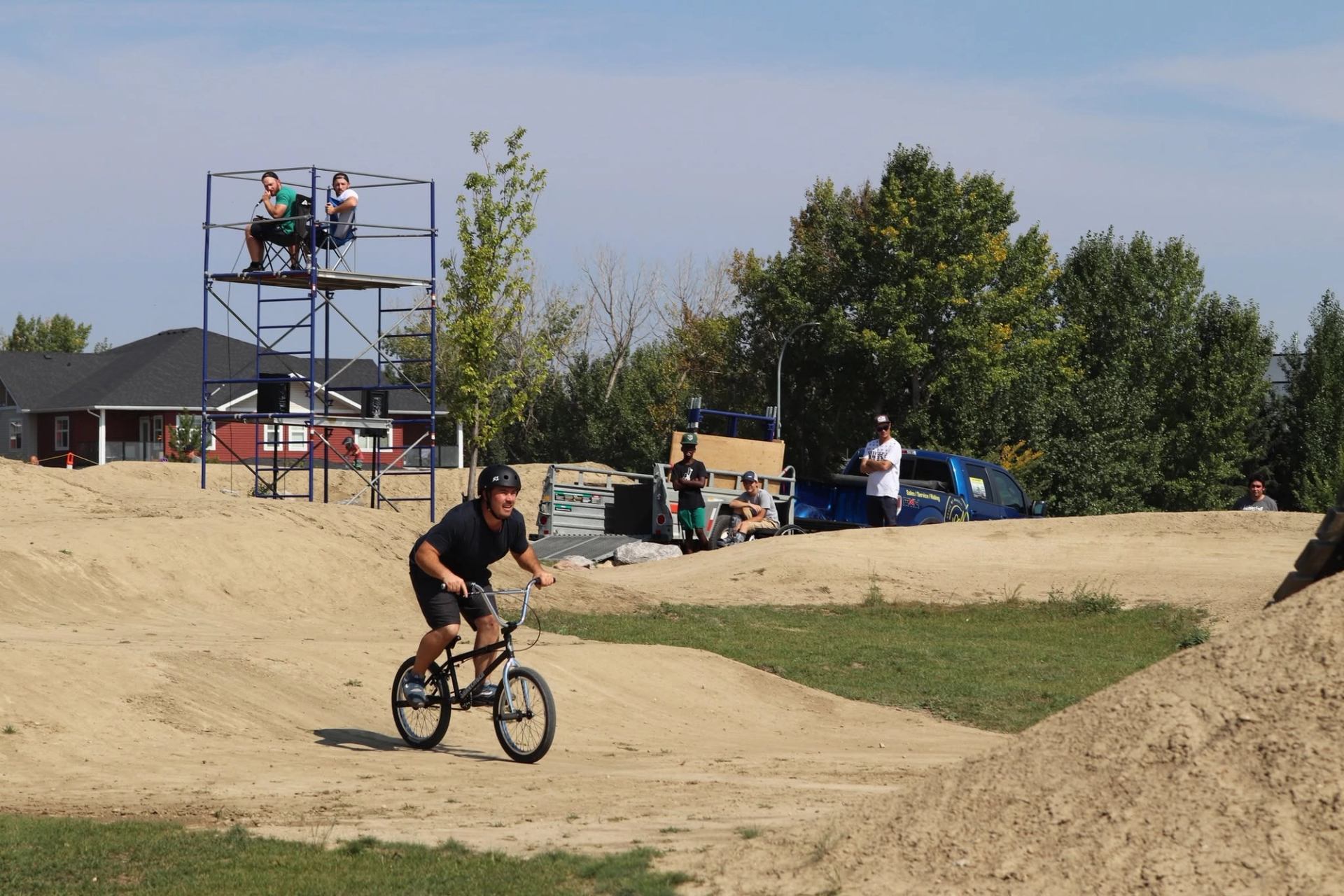 BMX rider pedals on dirt track while spectators watch from scaffolding and trucks.