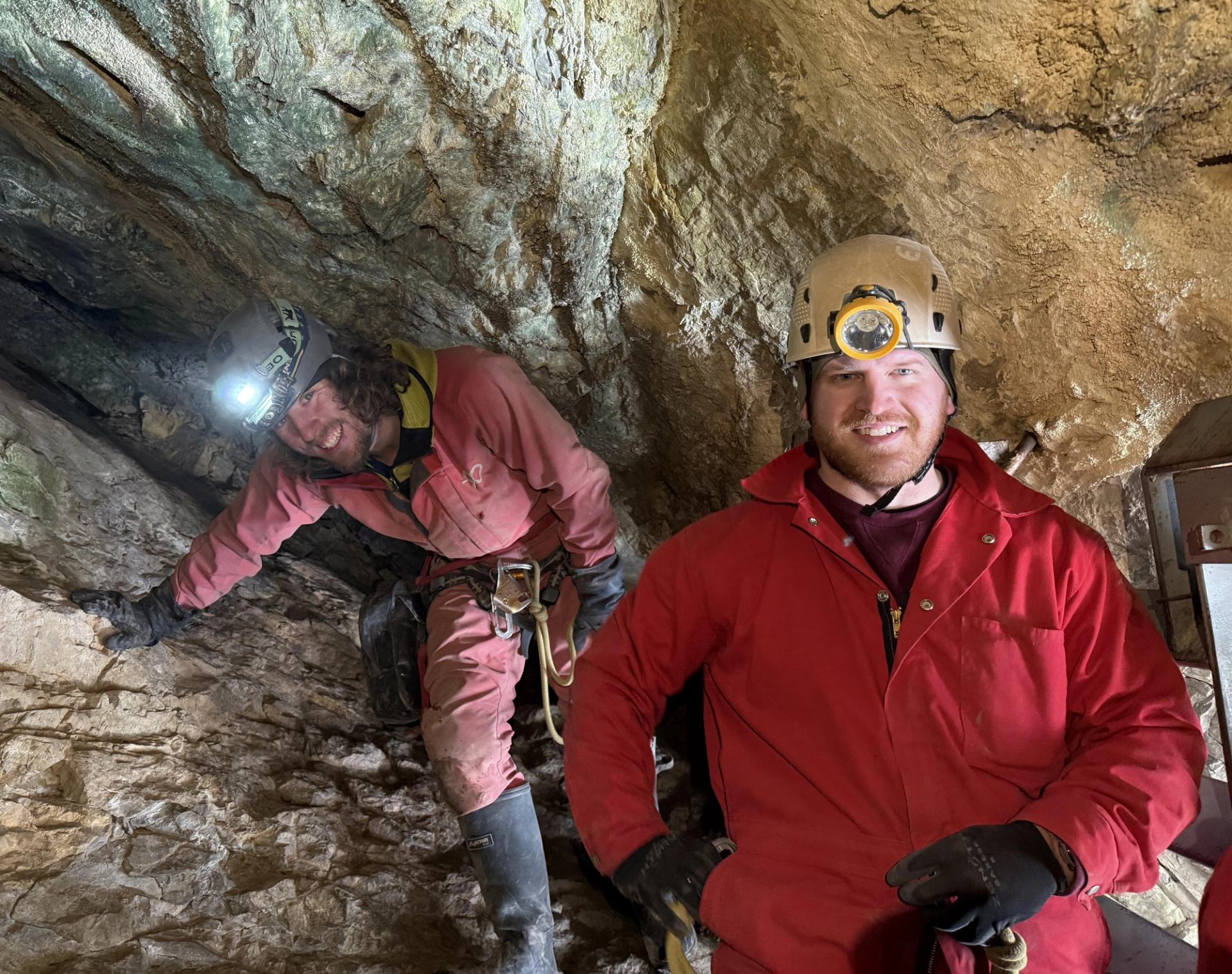 Cave explorers wearing coveralls, helmets and headlamps stand among rough rock walls.