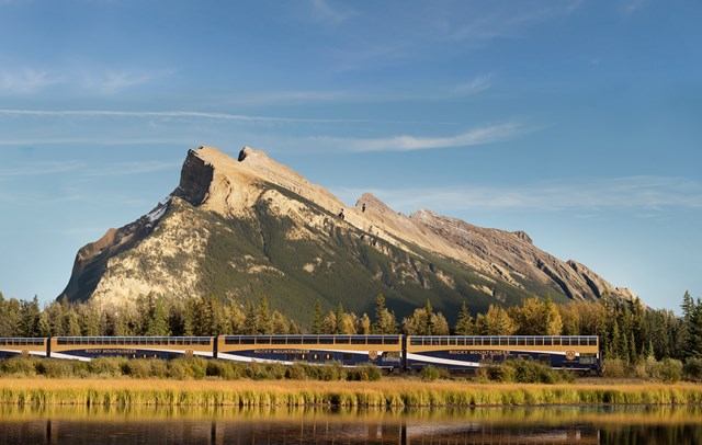 A train passes in front of a mountain beside a nearby lake.