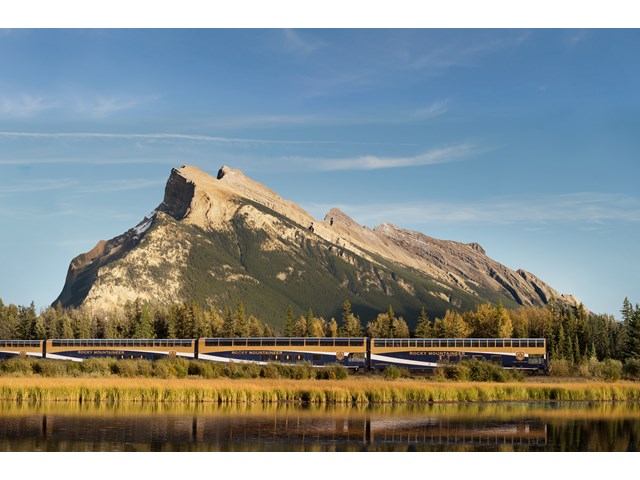 A train passes in front of a mountain beside a nearby lake.
