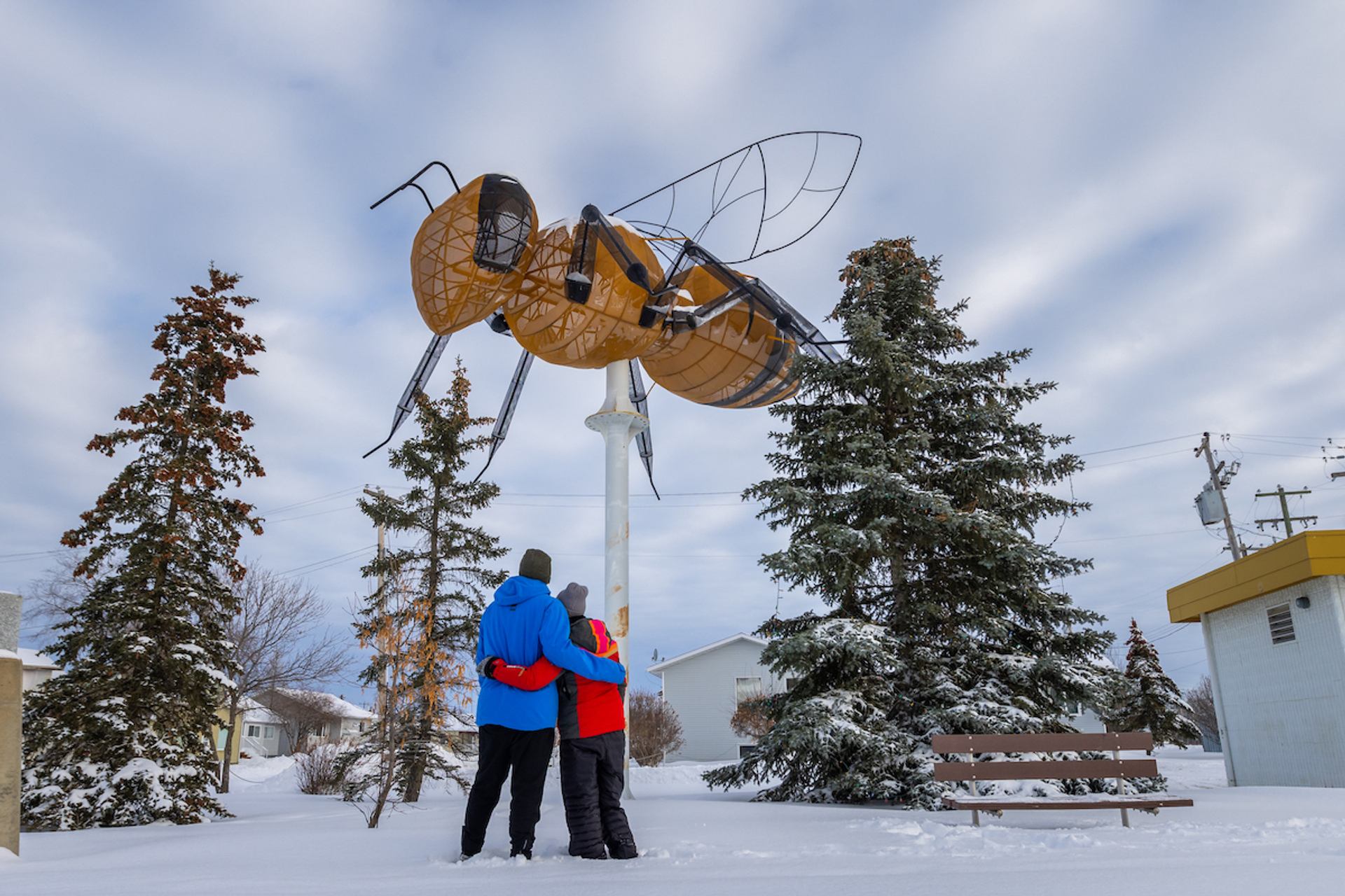 Two visitors stand beneath the World's Largest Bee sculpture surrounded by snow-covered trees. 