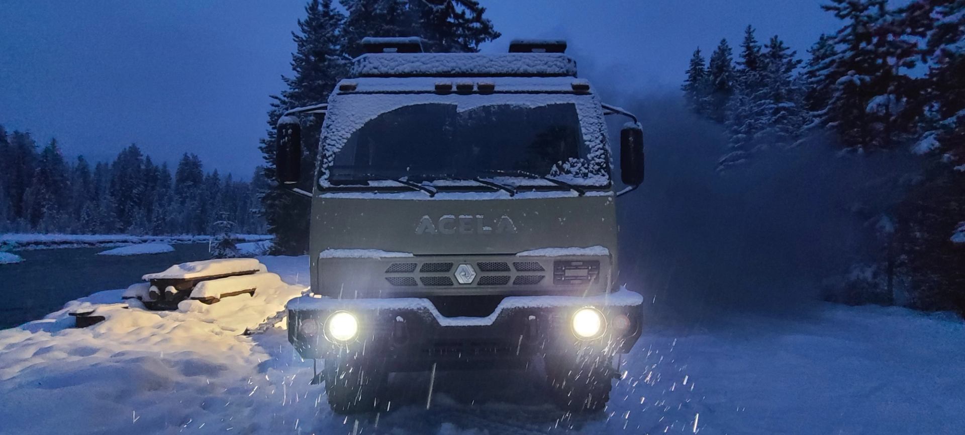 Acela expedition truck driving through snowy forest at dusk with headlights on.