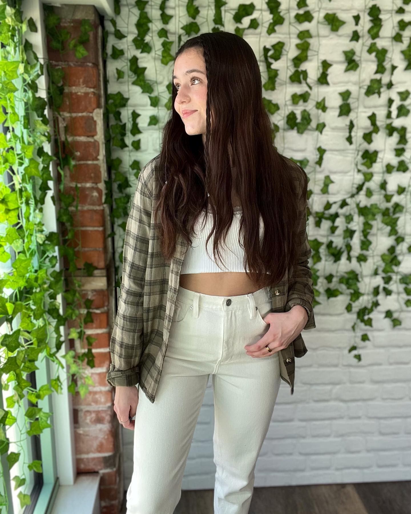 Woman standing in front of a green leaf wall and brick column, wearing casual clothing.