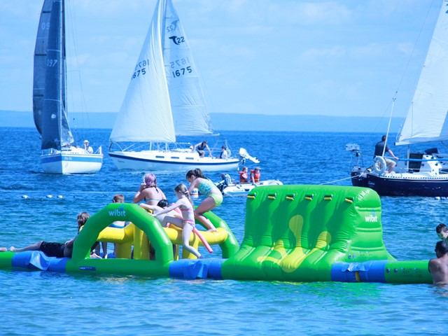 Kids enjoy a floating water obstacle course as sailboats cruise the bright blue lake in summer.