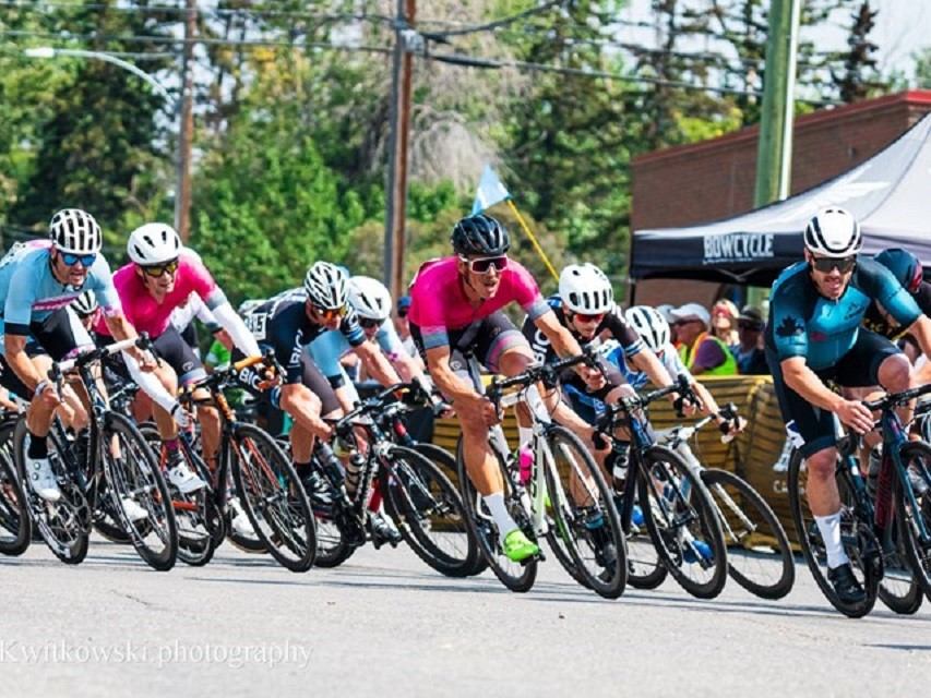 Cyclists in colourful jerseys race around a street corner past spectators and tents.