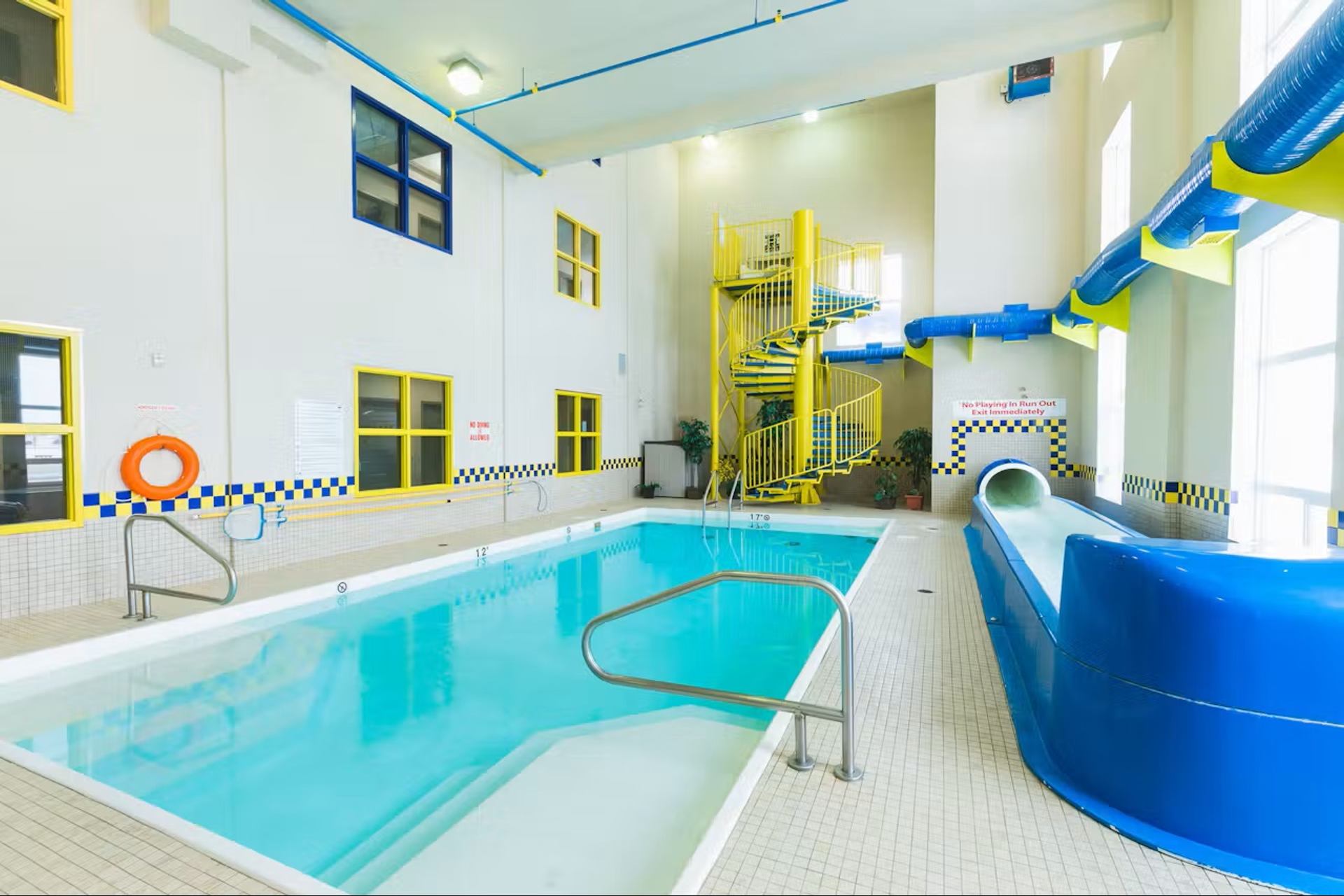 Indoor pool with turquoise water, blue waterslide, spiral staircase, and bright sunlit windows.