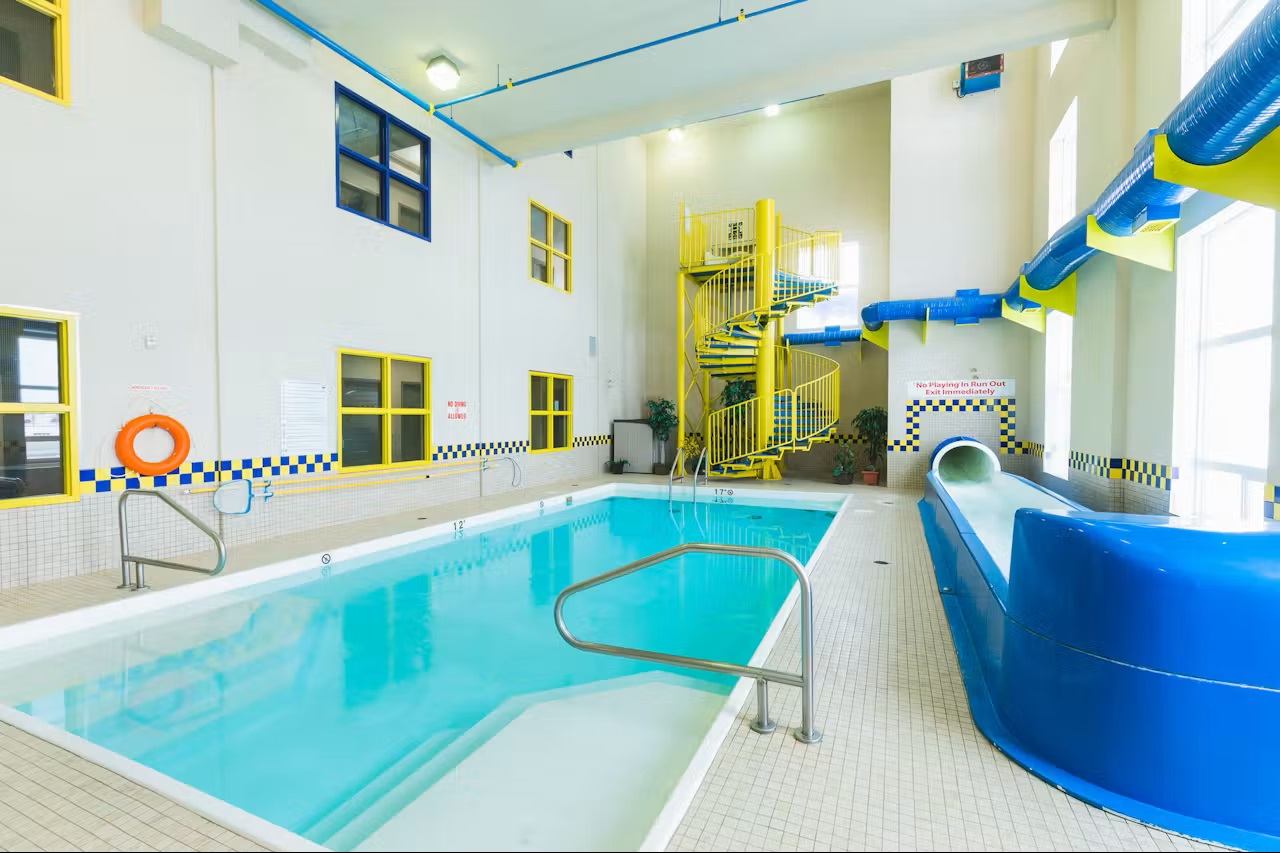 Indoor pool with turquoise water, blue waterslide, spiral staircase, and bright sunlit windows.