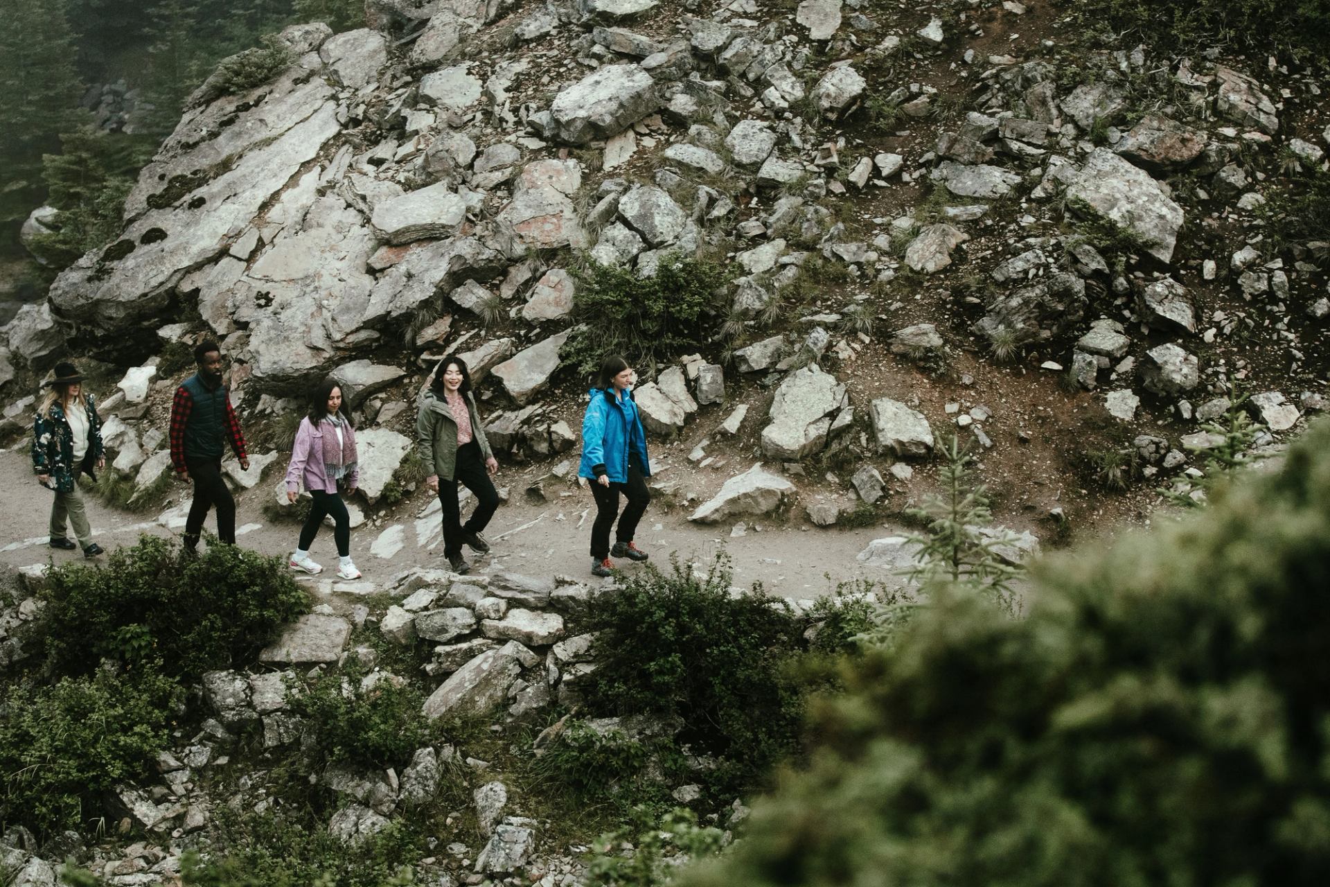Hikers walk along a rocky mountain trail bordered by shrubs and evergreen trees.