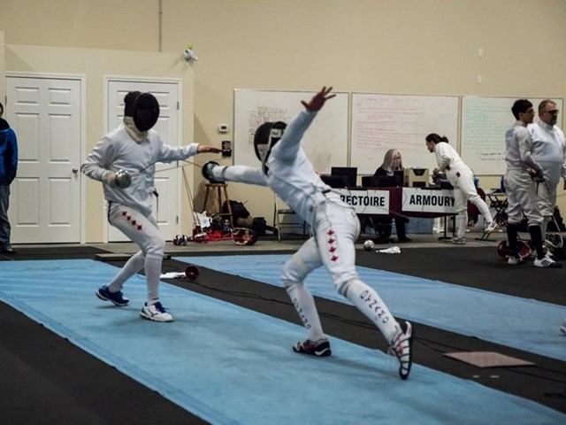 Fencers in white fencing gear practicing on a blue piste in a sports room.