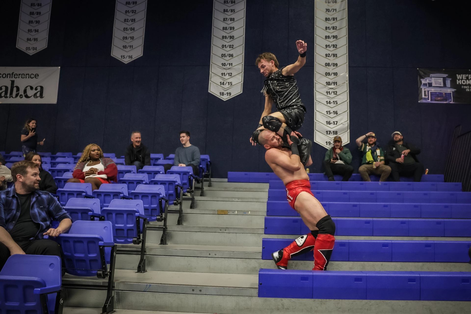 Professional wrestlers fight on arena steps as scattered spectators watch from blue seats.