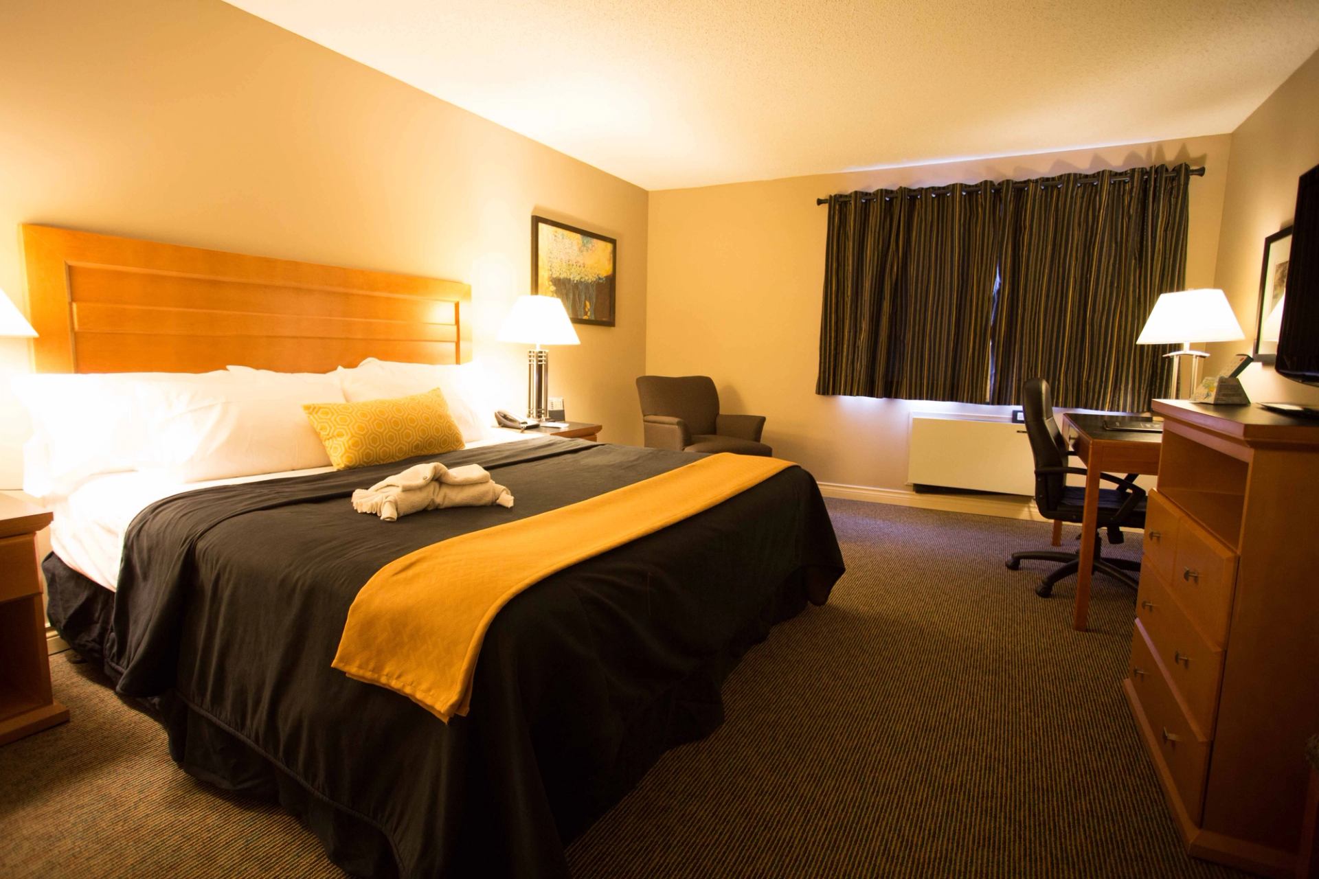 A white towel folded into an elephant shape sits on a neatly made bed with black and mustard bedding. A geometric mustard pillow adds contrast. Cozy ambiance.