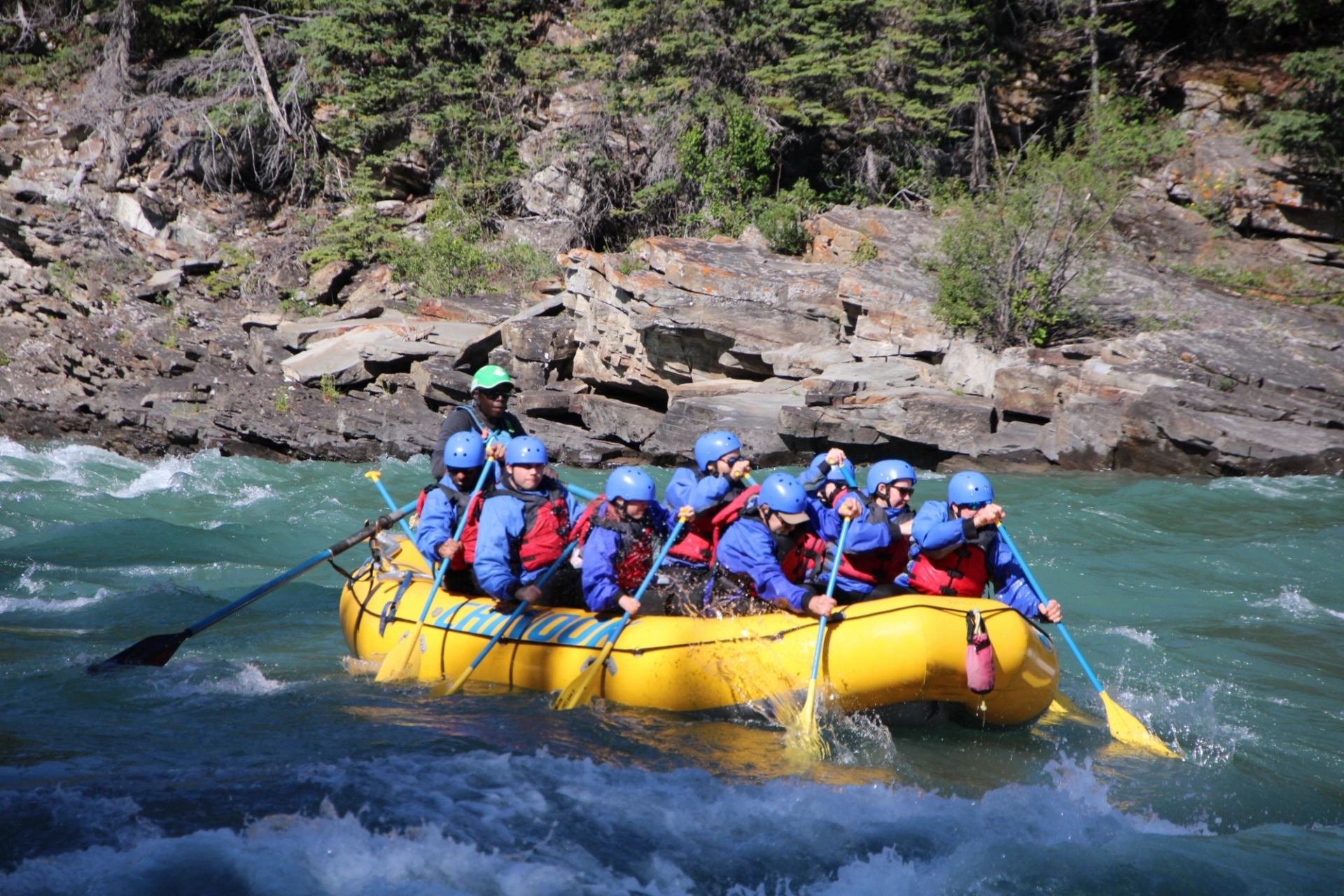 Group in helmets paddles yellow raft through river rapids beside rocky forested shore.