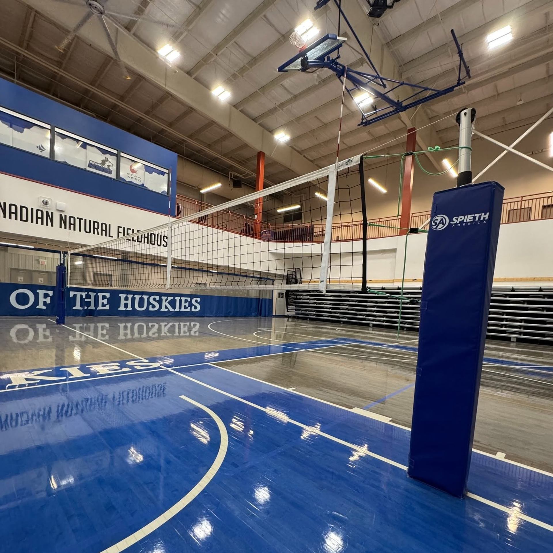 Indoor volleyball court with net, blue floor markings and empty spectator bleachers.