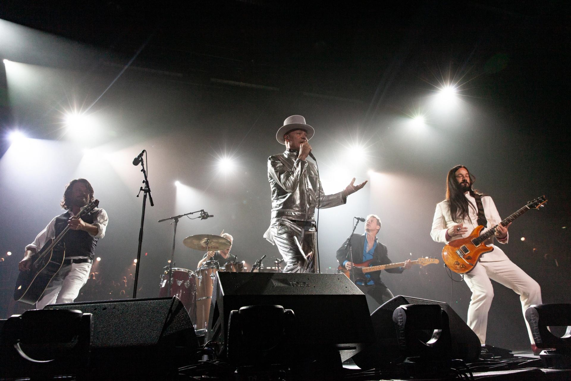 Rock band performs on stage under bright spotlights, with singer in metallic suit and hat.