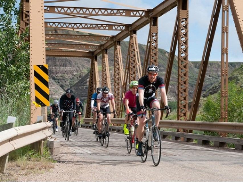 Cyclists ride across a metal truss bridge with rocky hills and greenery in background.