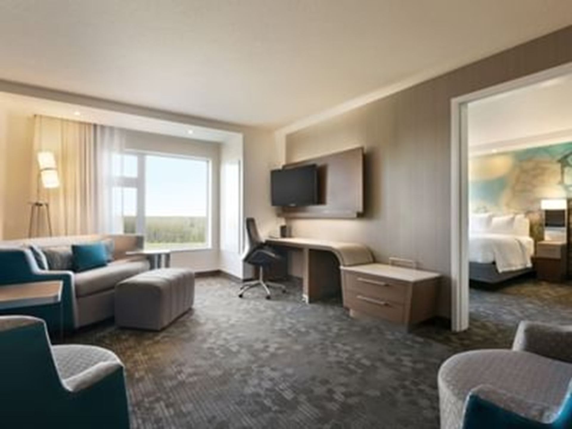 Modern hotel suite with seating area, desk, wall-mounted TV and adjoining bedroom.