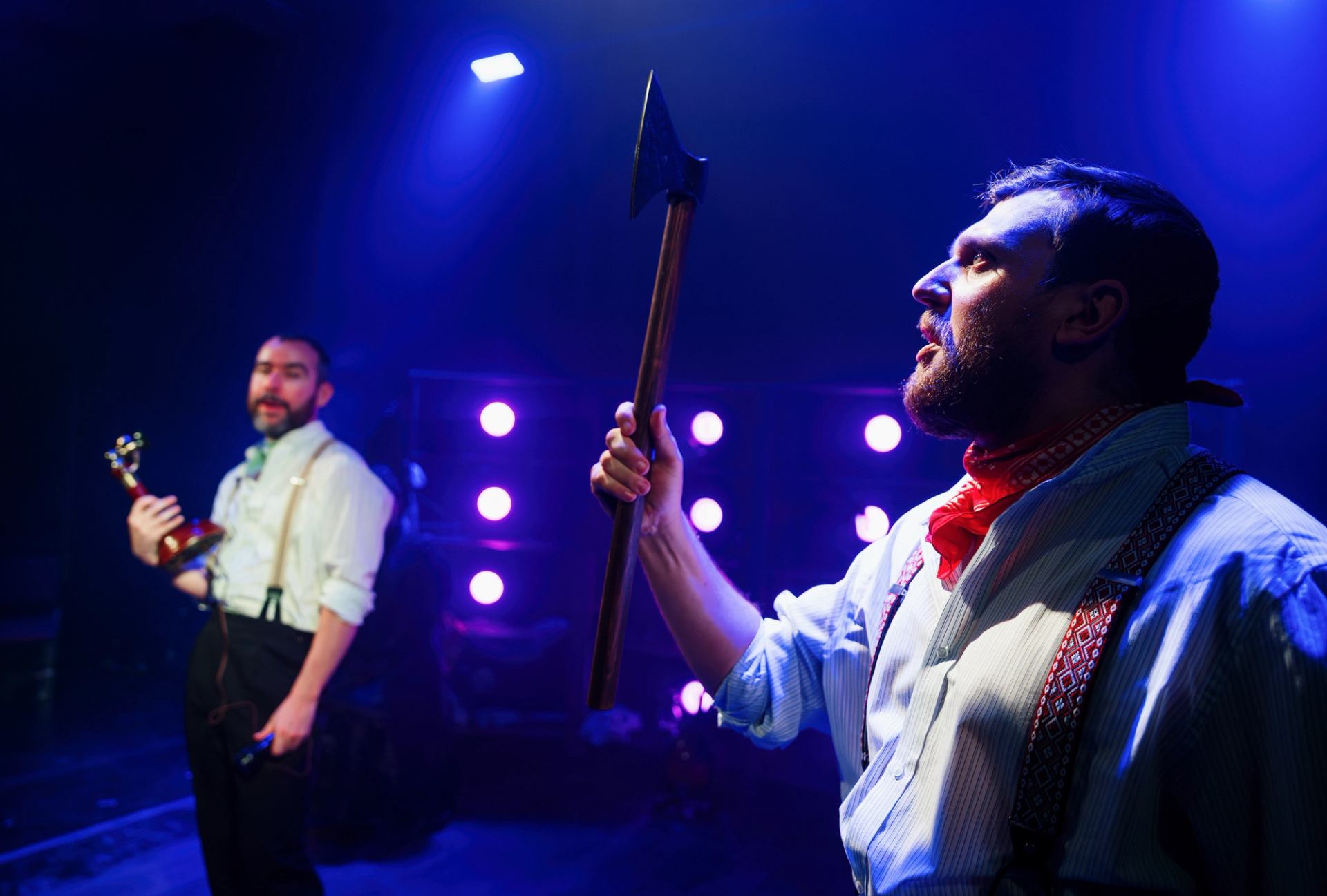 Stage performer holds an axe under blue lights as another holds a trophy behind.