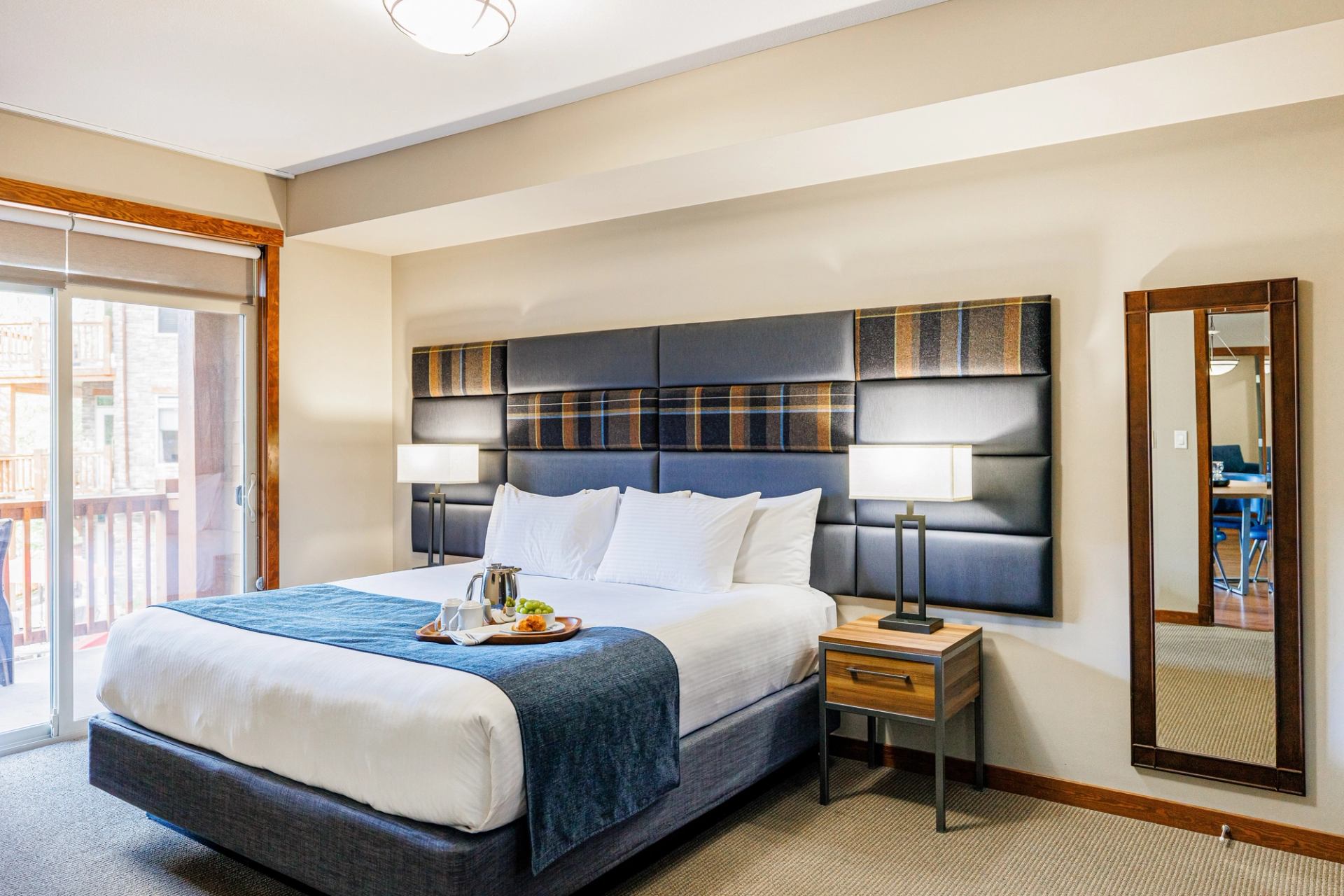 A cozy hotel room with a bed, food tray, modern lamp, and a view through a large window.