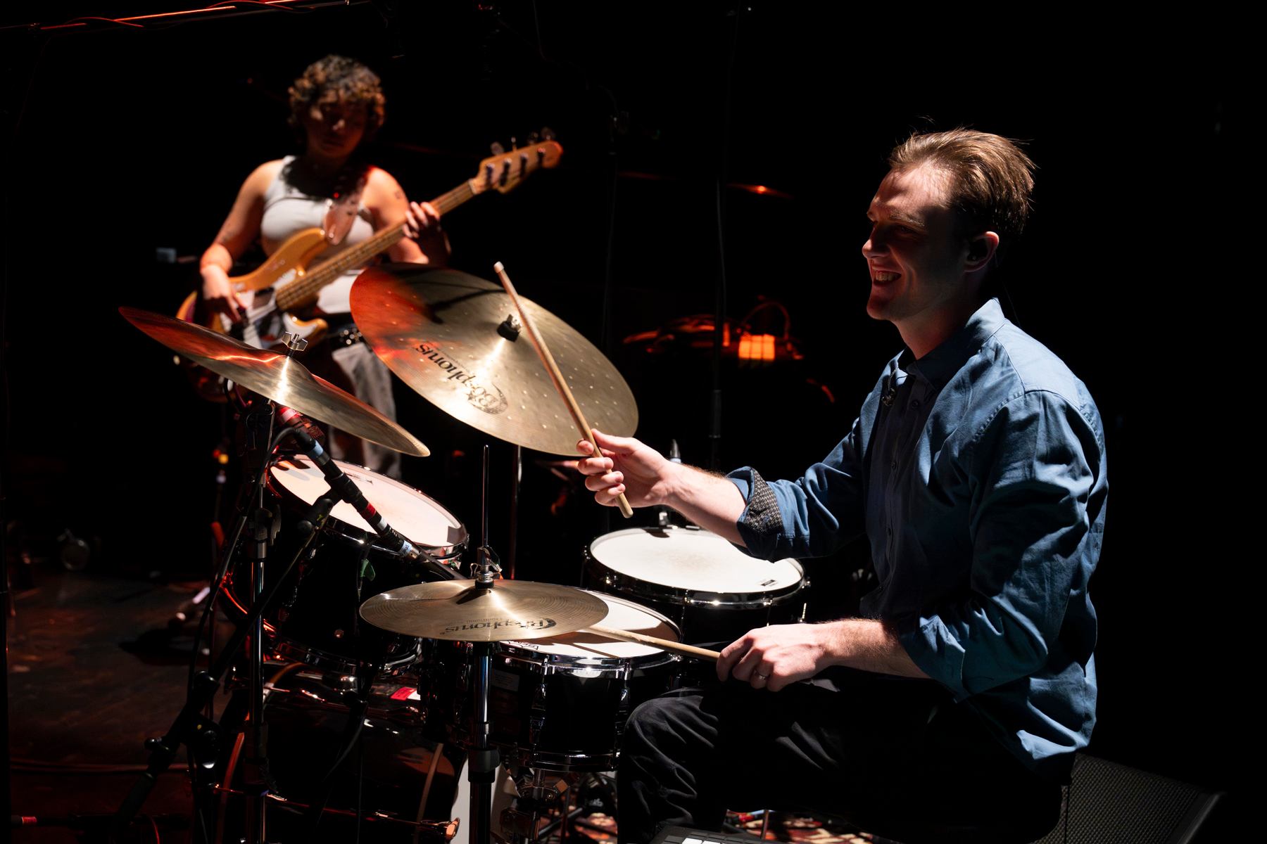 Drummer plays a drum kit onstage while a bassist performs in the background.
