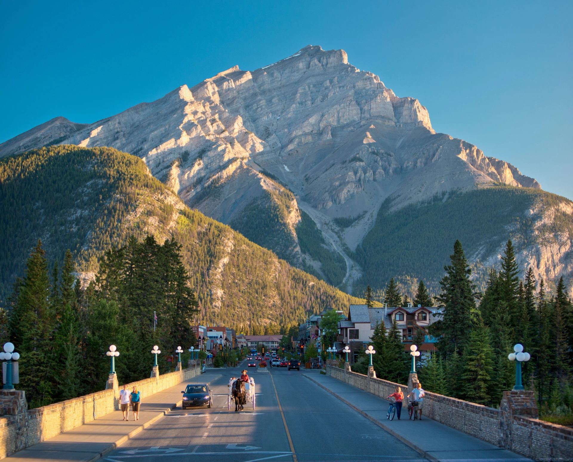 Mountain towering over a small town street with cars, pedestrians and evergreen trees.