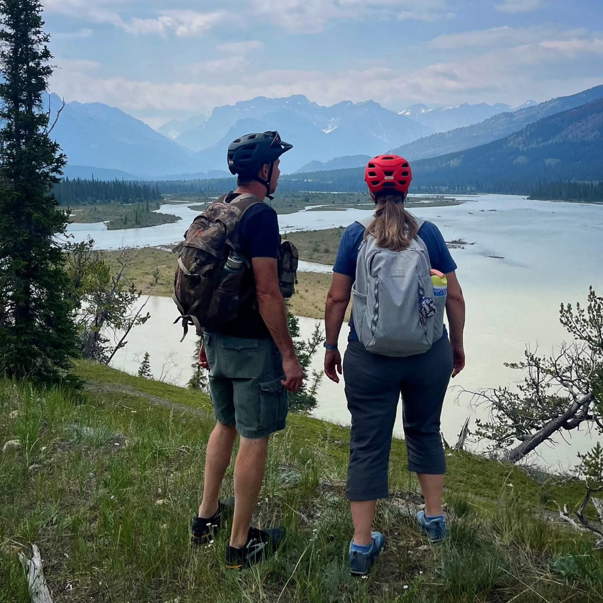 Cyclists with backpacks stand on grassy hill overlooking winding river and distant mountains.