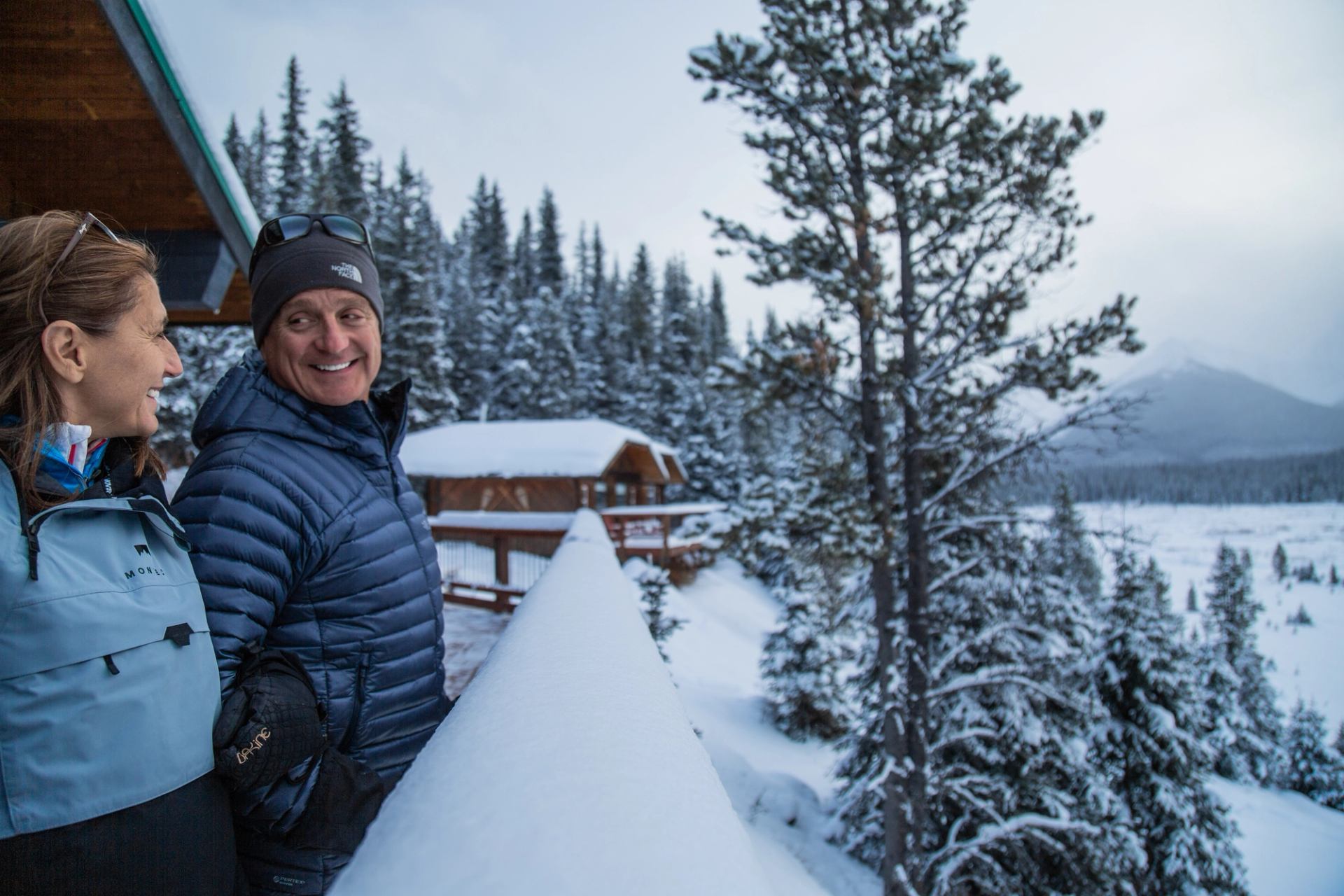 Two people standing on a lodge balcony overlooking snow-covered trees and distant peaks