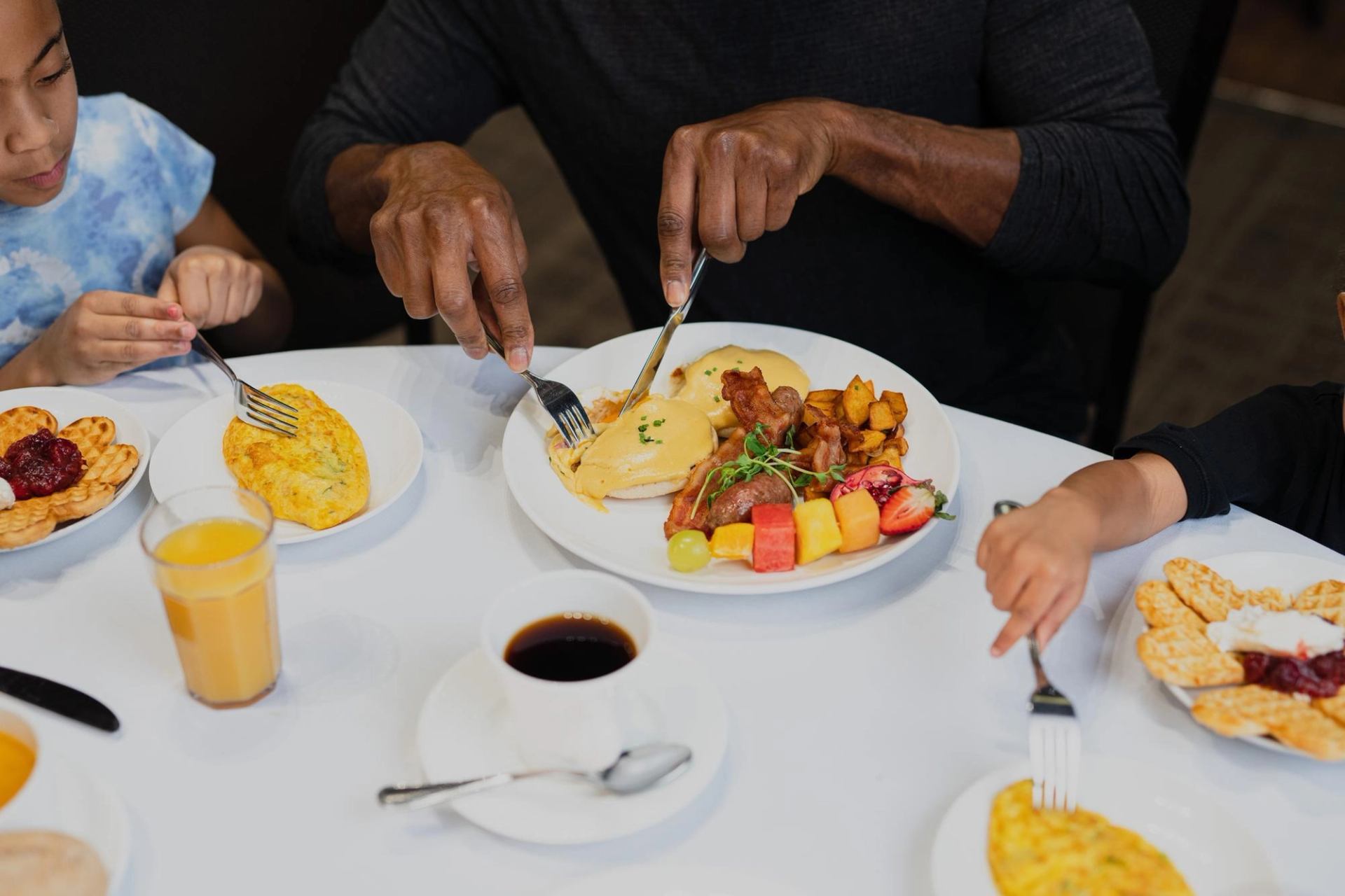 A group eats breakfast at a white table with Eggs Benedict, omelets, waffles, fruit, coffee, and juice.
