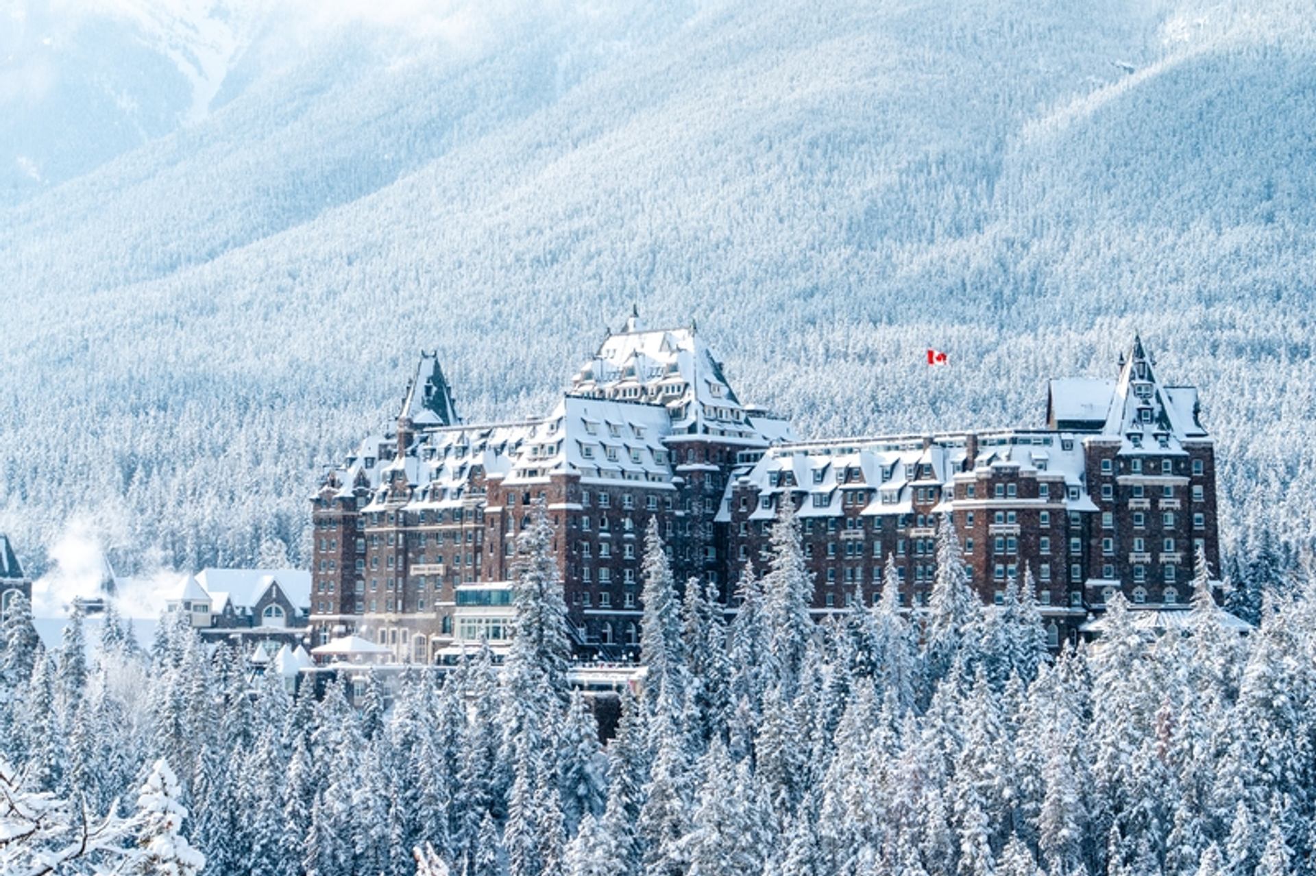 Fairmont Banff Springs stands above snow-covered evergreen forest, framed by mountain slopes in winter.
