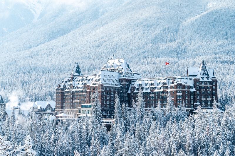 Fairmont Banff Springs stands above snow-covered evergreen forest, framed by mountain slopes in winter.