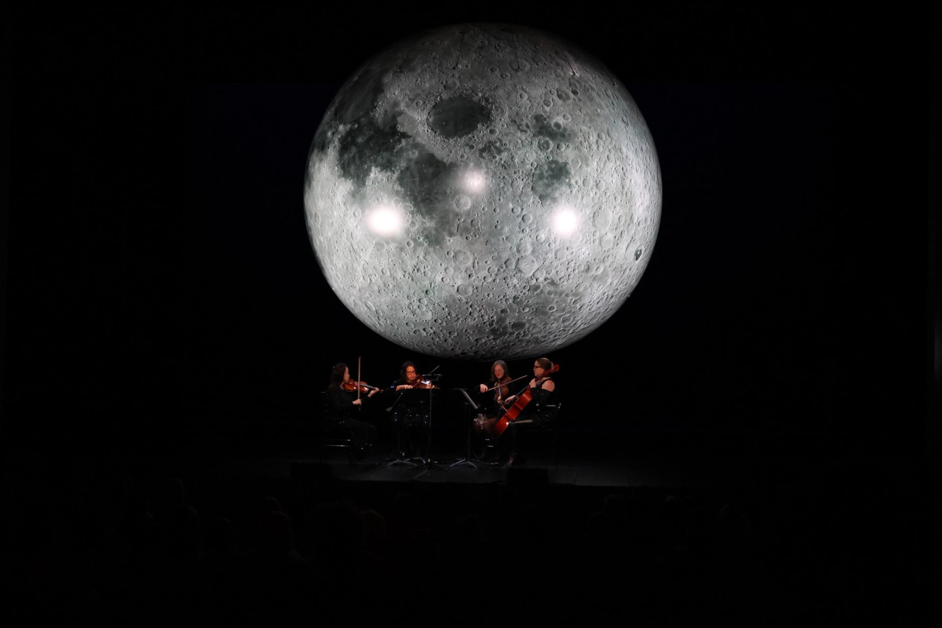 String quartet performs on dark stage beneath large illuminated moon projection backdrop.