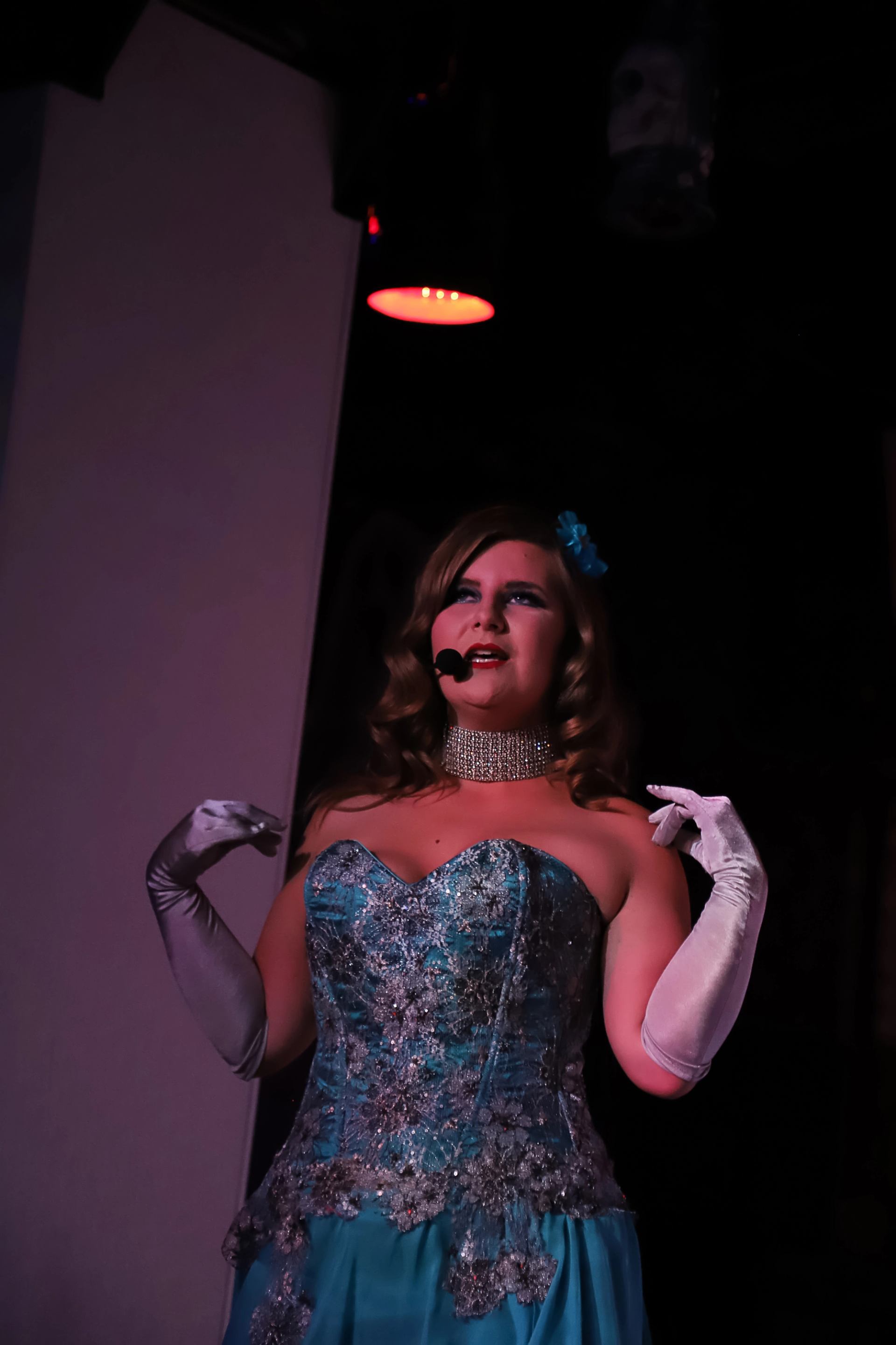 A woman performs in a sparkly blue corset dress, long white gloves, a rhinestone choker, and a headset microphone.