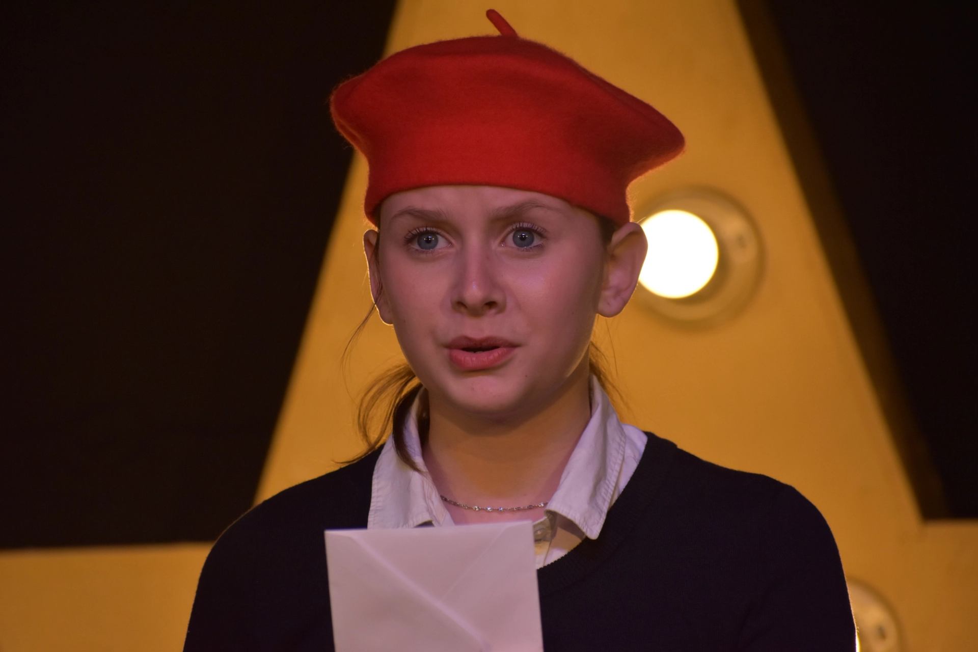 Person in red beret holds paper near a lit letter "A" on stage.