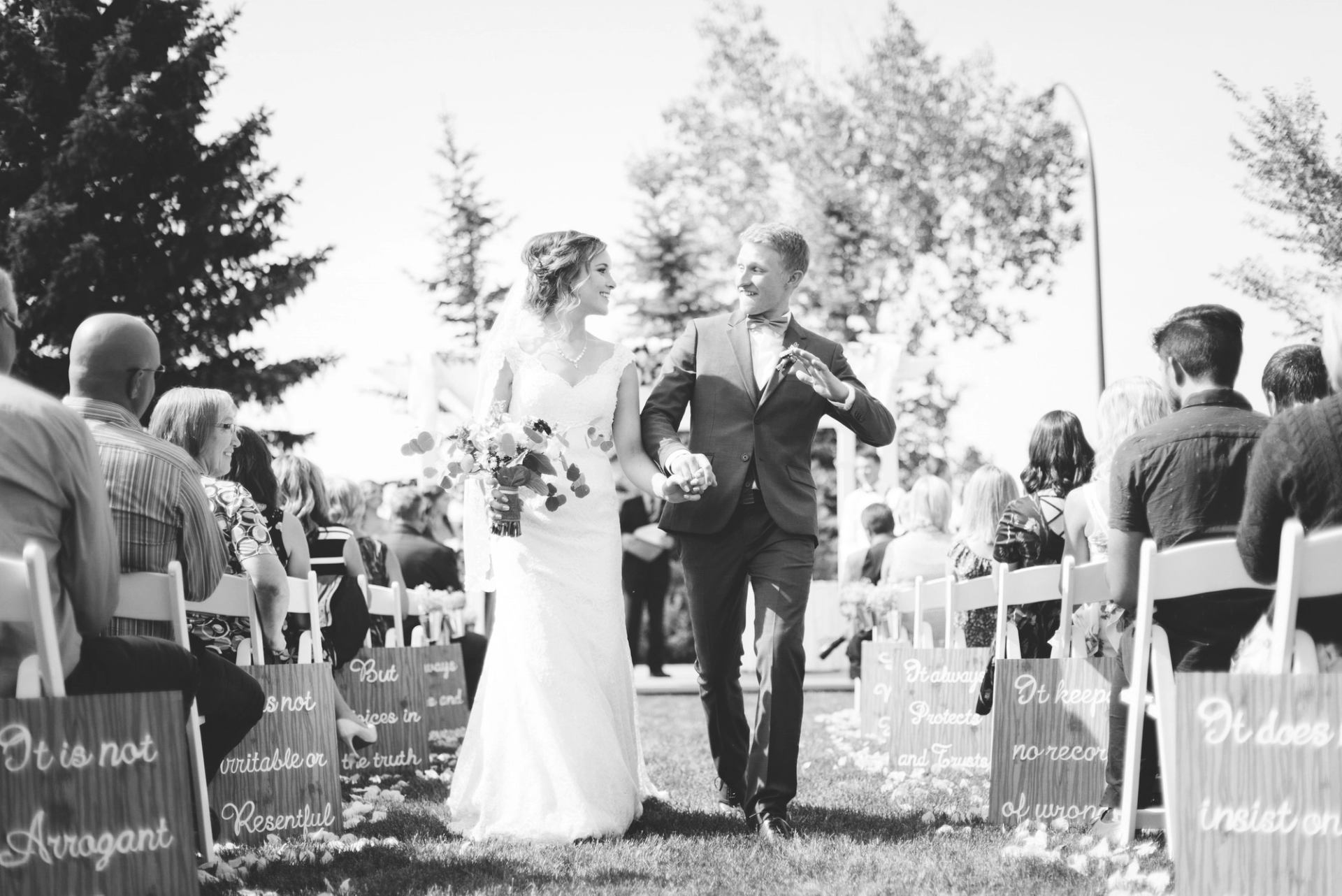 Newlyweds walk hand in hand down an outdoor aisle lined with seated guests.