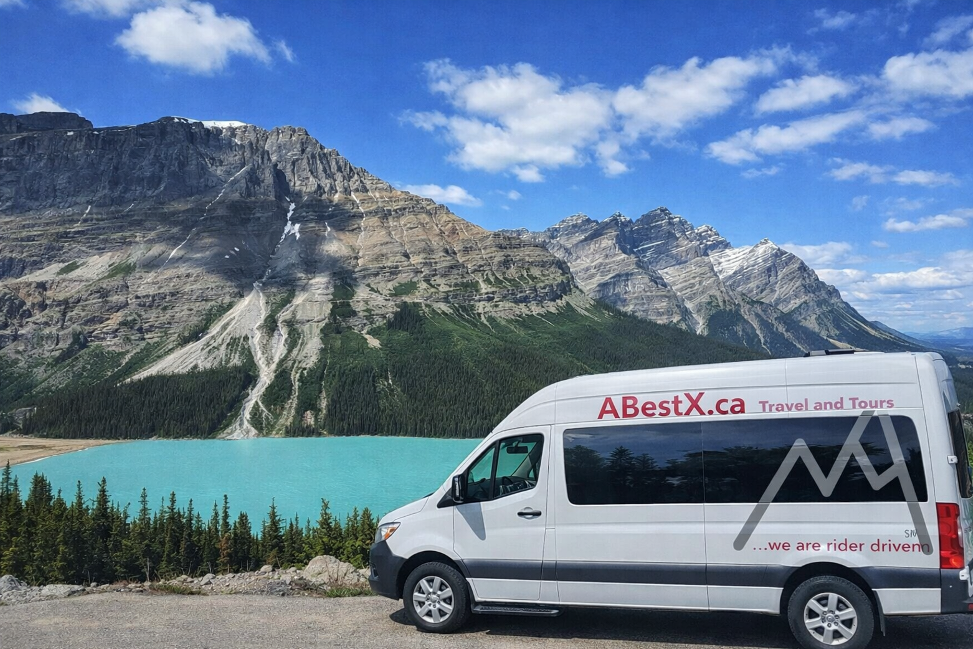 Tour van marked ABestX.ca parked beside turquoise lake and rocky mountain range.