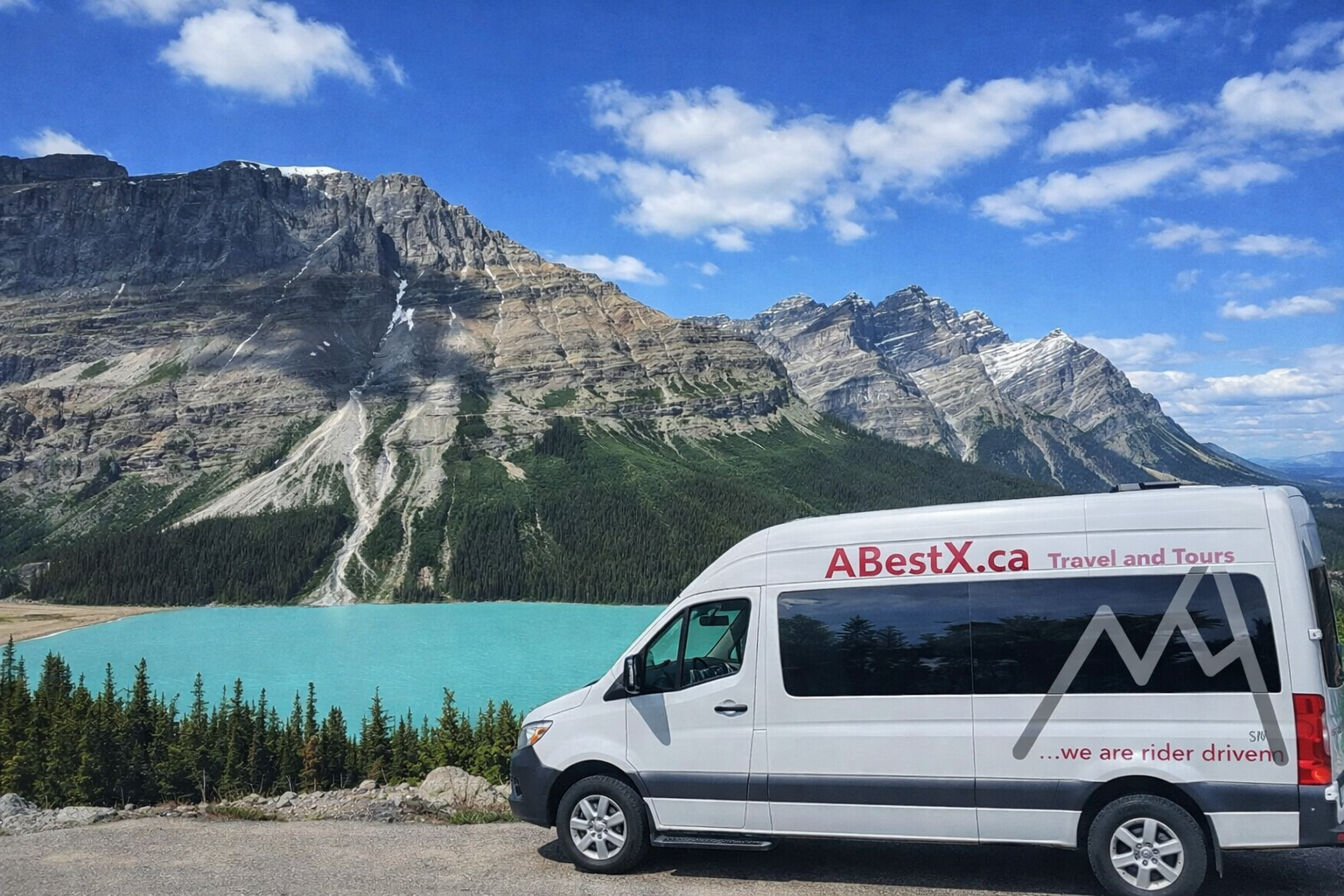 Tour van marked ABestX.ca parked beside turquoise lake and rocky mountain range.