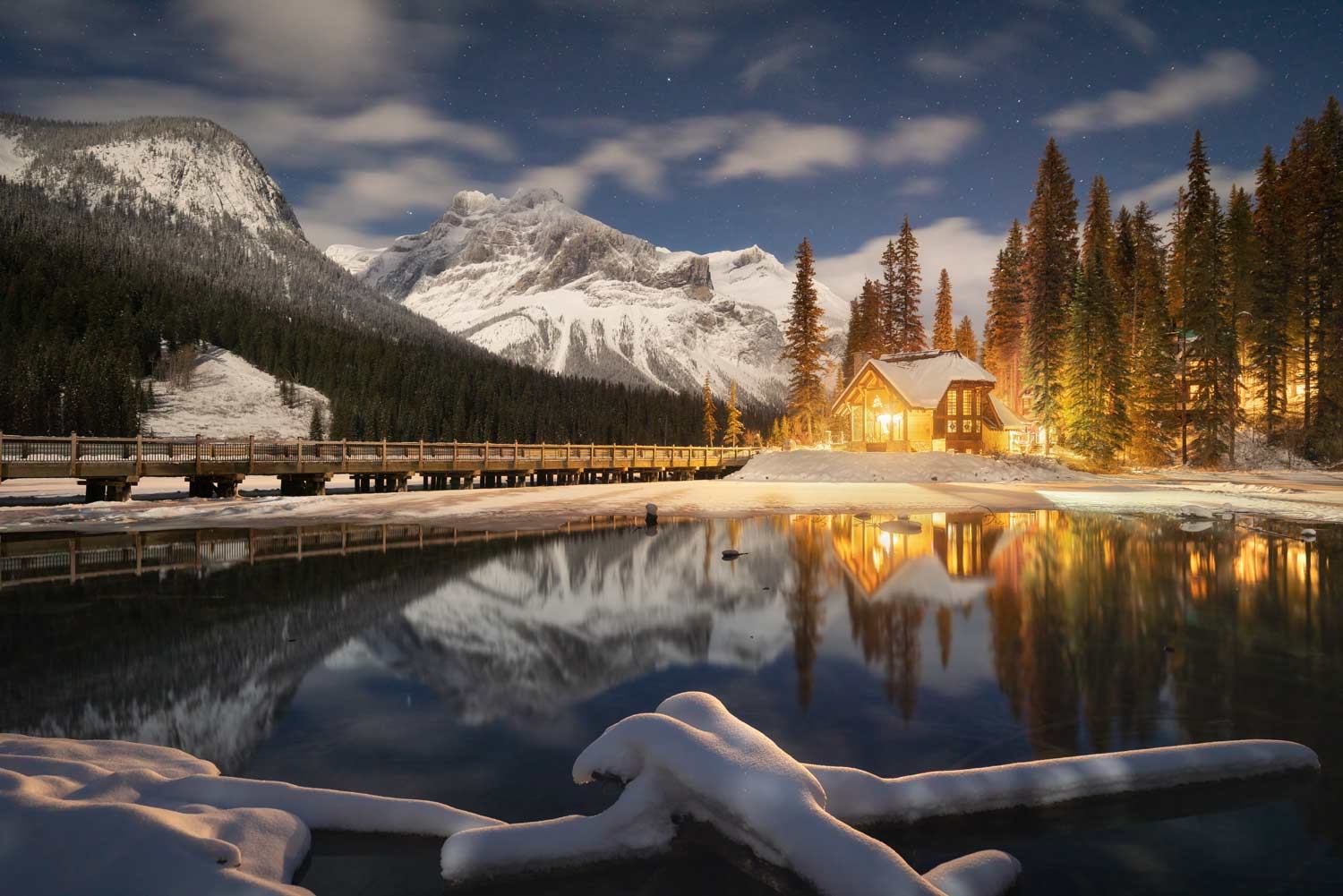 A cozy cabin glows by a reflective, partially frozen lake in a snowy mountain landscape at night, with a long wooden boardwalk and a starry sky.