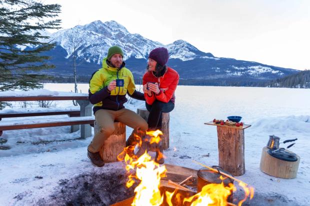 A couple relax and enjoy hot drinks around an open campfire at Pyramid Lake in Jasper National Park.