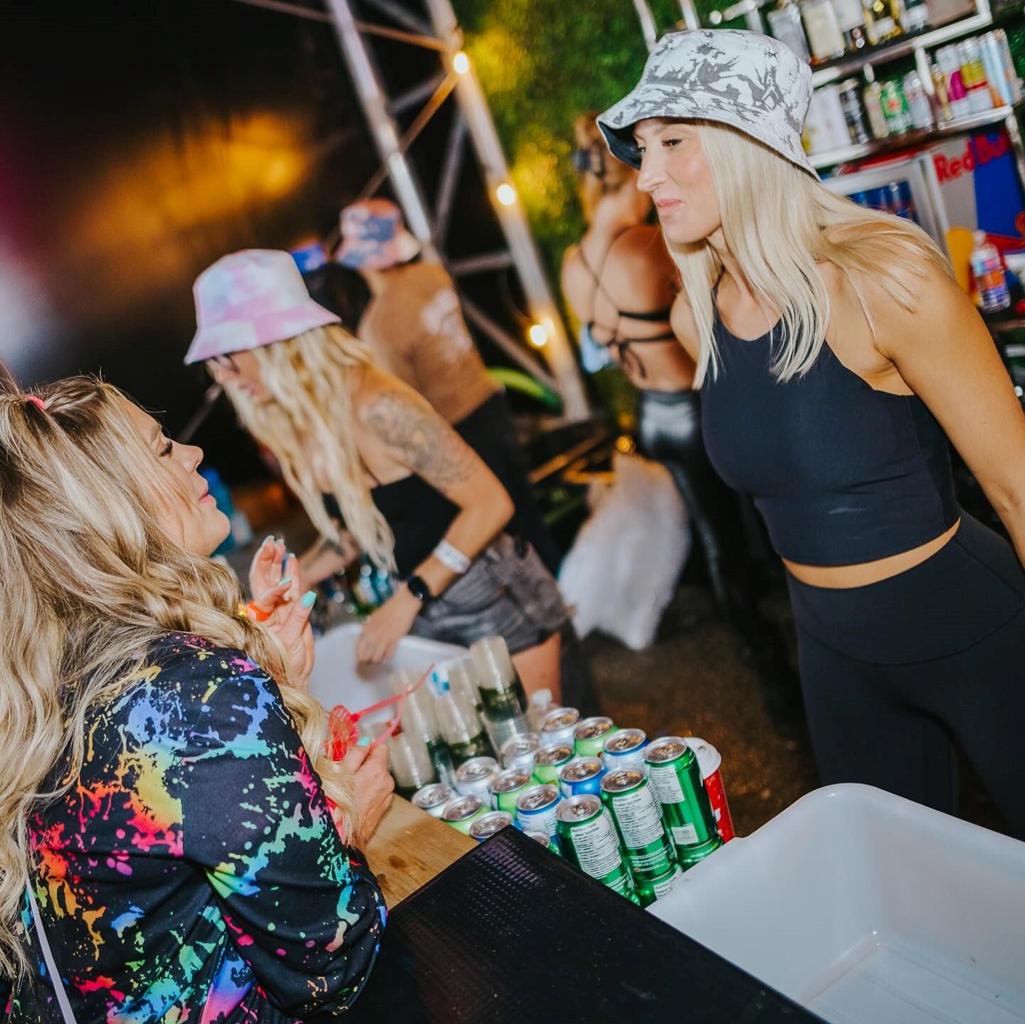 Festival attendees at a table with drinks cans and outdoor lights, night concert setting.