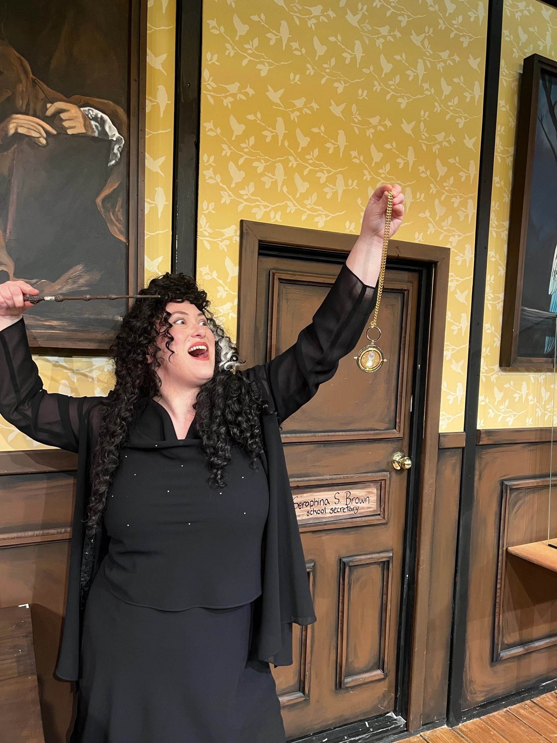 Person in black costume holds wand and pocket watch on stage with painted backdrop.
