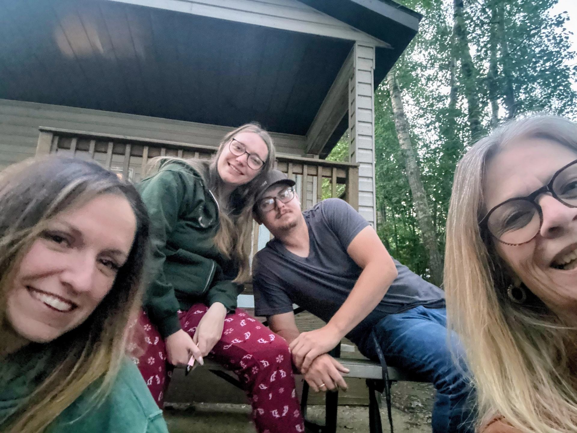 Four people sit on a cabin porch bench, leaning toward the camera.