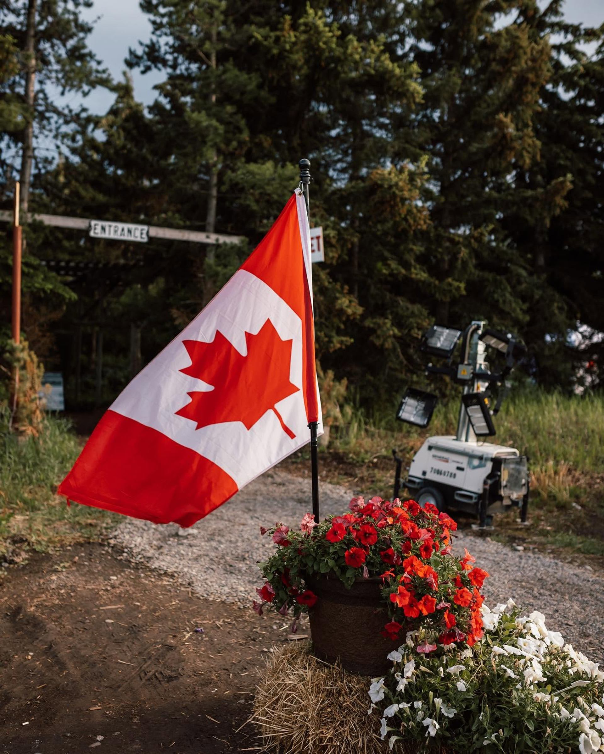 Canadian flag beside red and white flowers on hay bale near forest entrance.
