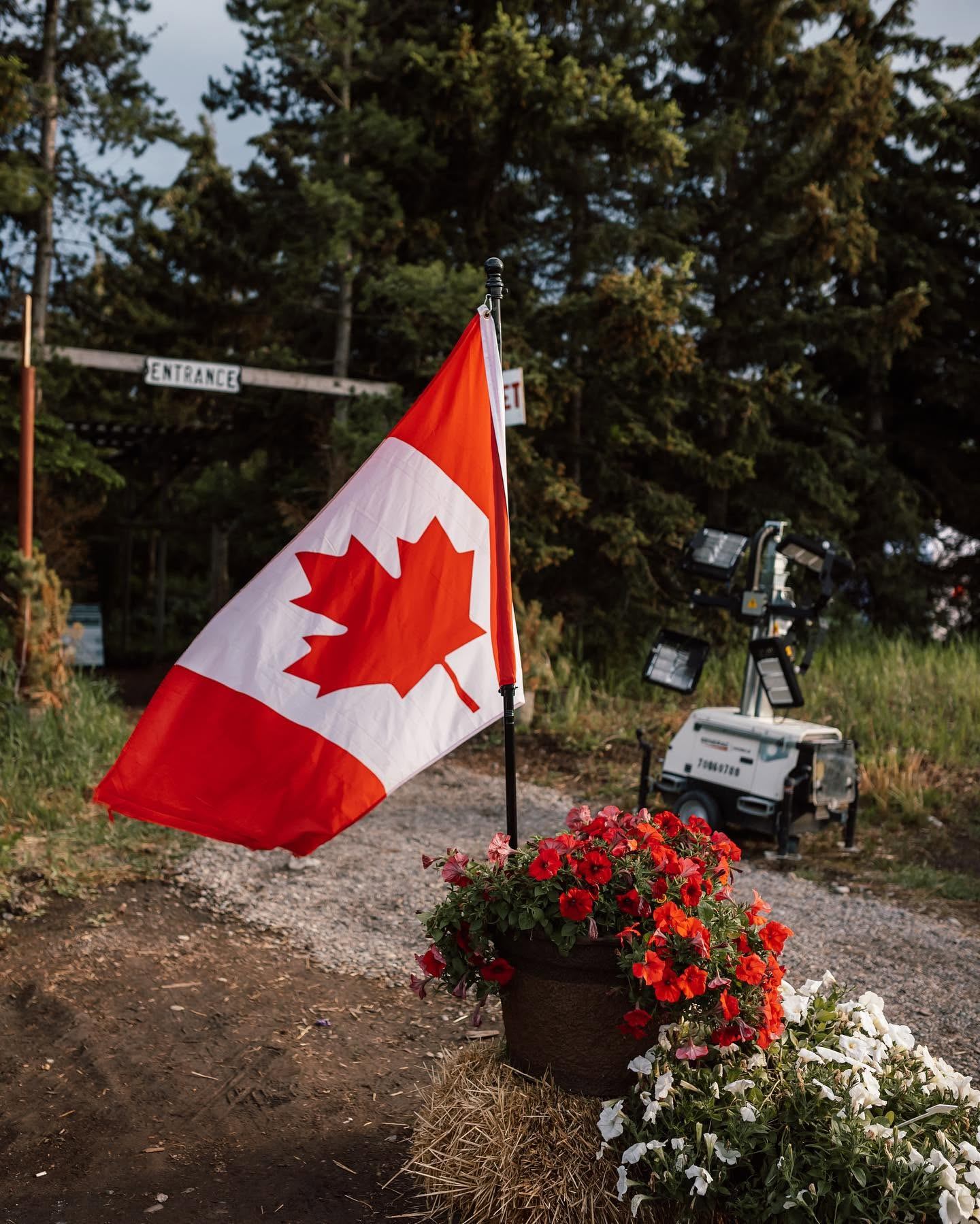 Canadian flag beside red and white flowers on hay bale near forest entrance.