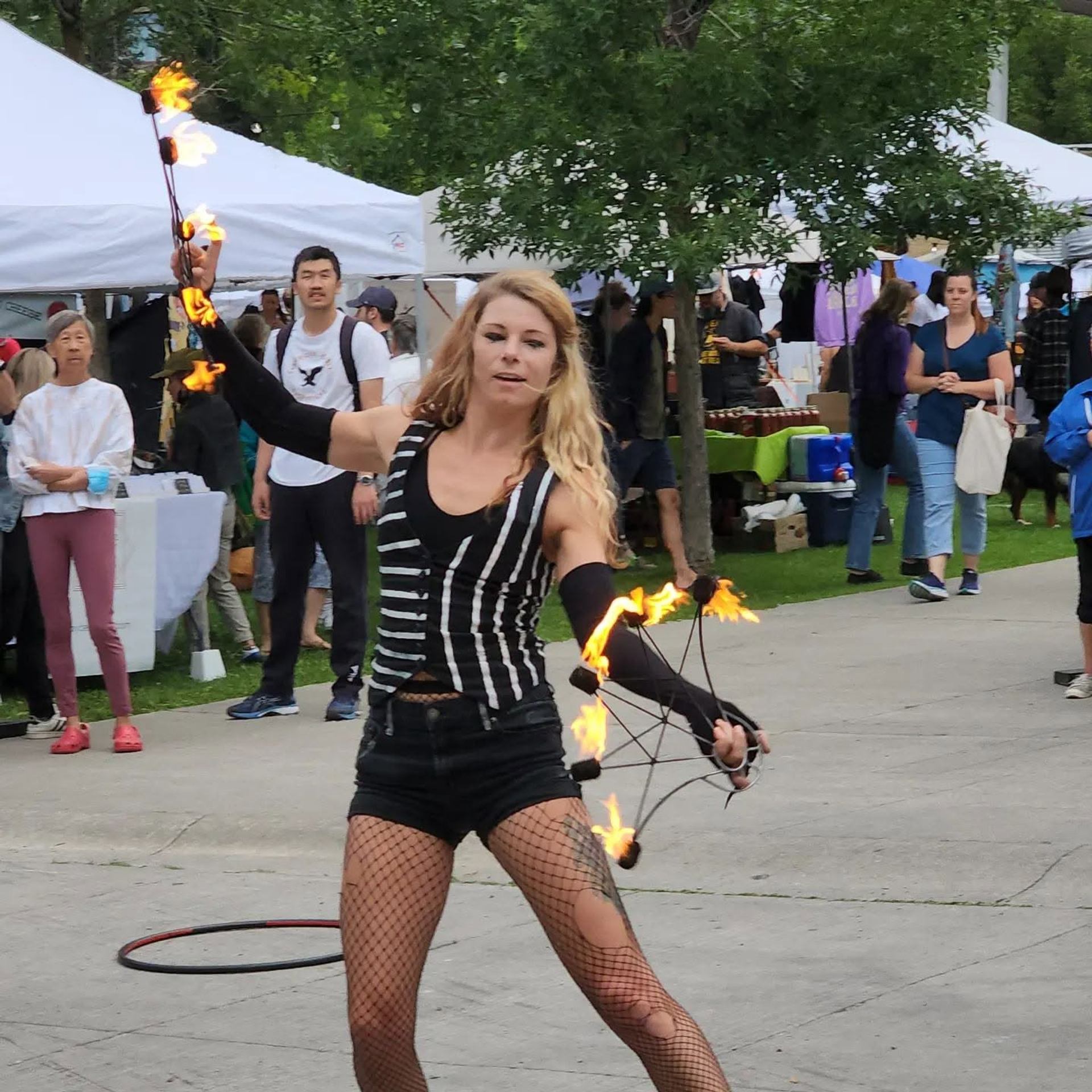 Street performer spins flaming props at outdoor market while people watch in background.