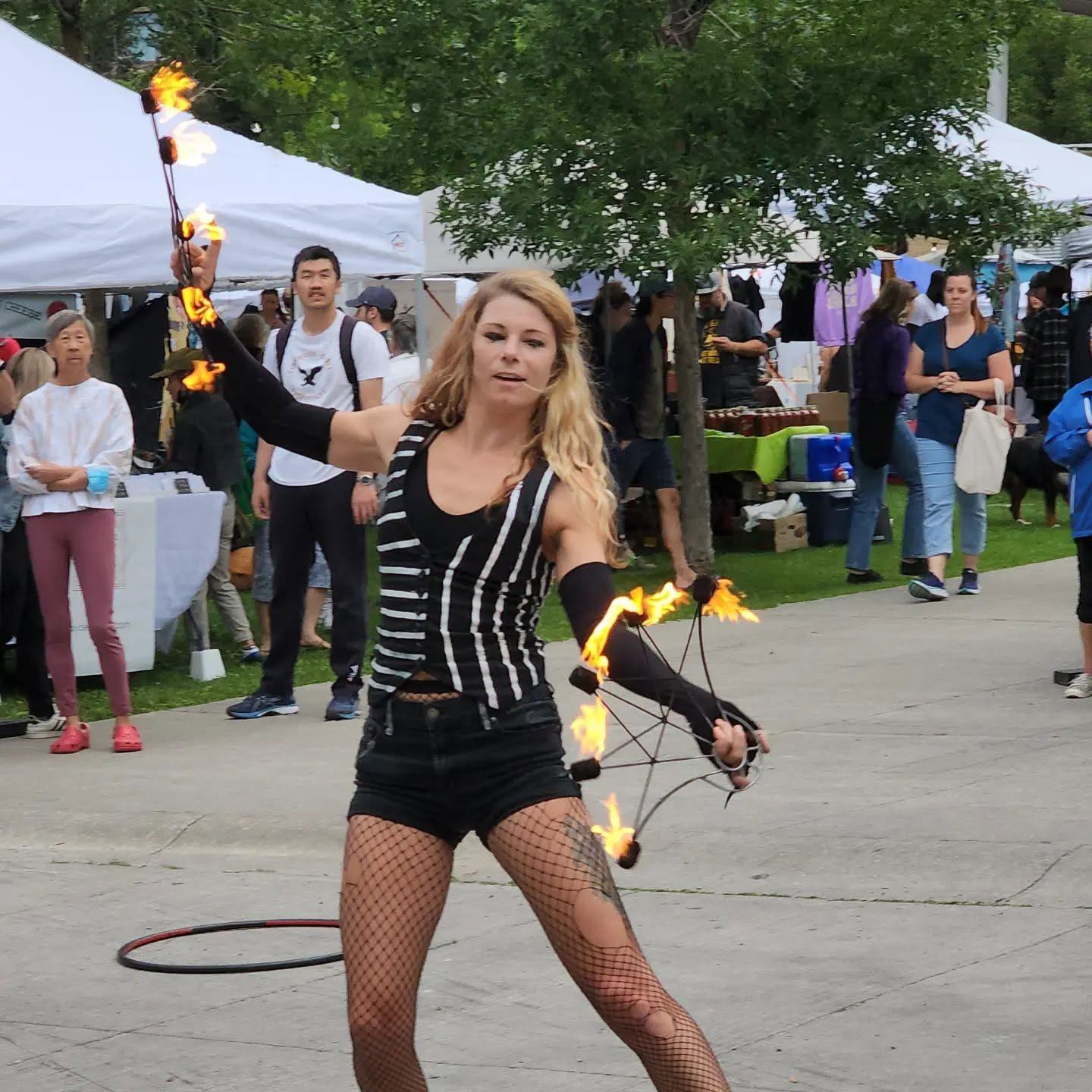 Street performer spins flaming props at outdoor market while people watch in background.