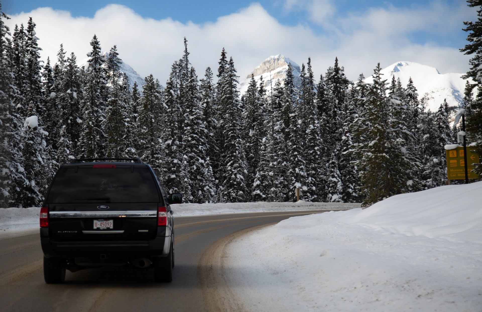 Black SUV driving along a snow-lined road surrounded by tall pine trees and mountain peaks.