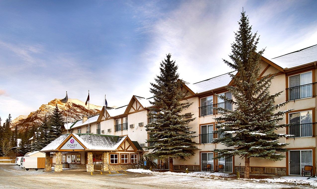 Rustic hotel exterior with stone and wood, set against snow-capped mountains.